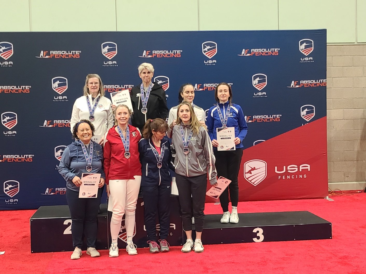 Group of female athletes standing on a winners' podium holding certificates and medals at a fencing competition. The backdrop features the USA Fencing logo and branding.