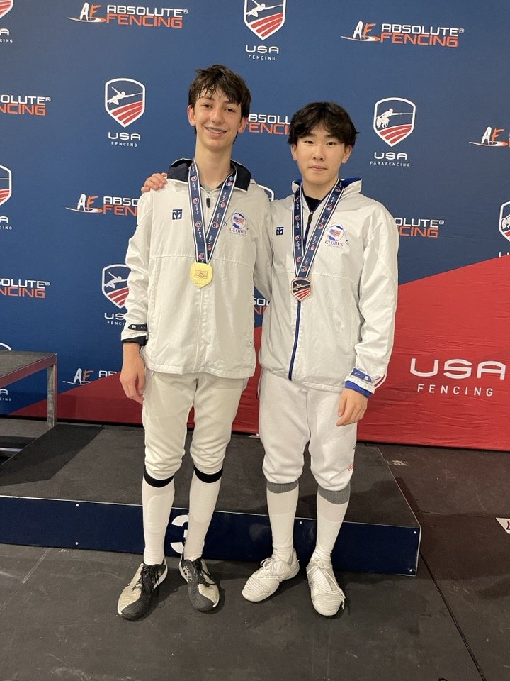 Two young male athletes in white fencing uniforms with medals around their necks, standing on a podium at a fencing competition. The backdrop features logos of USA fencing and Absolute Fencing.
