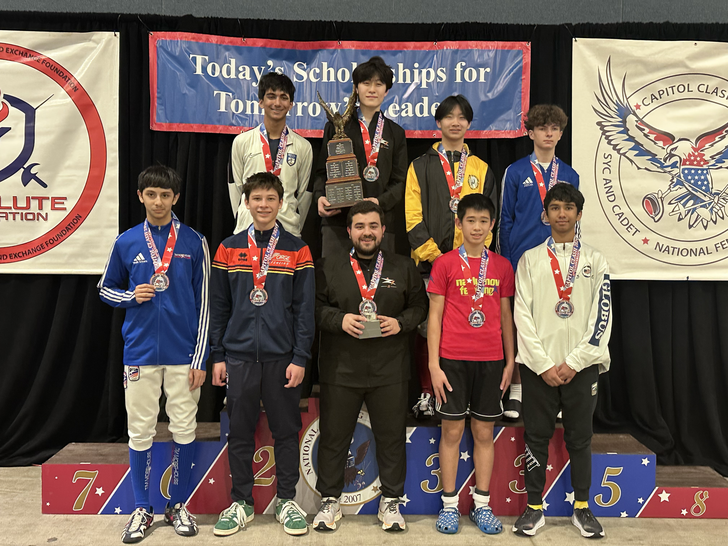 Young male fencers standing on a winners' podium, wearing medals and holding trophies, with a backdrop indicating a fencing competition.
