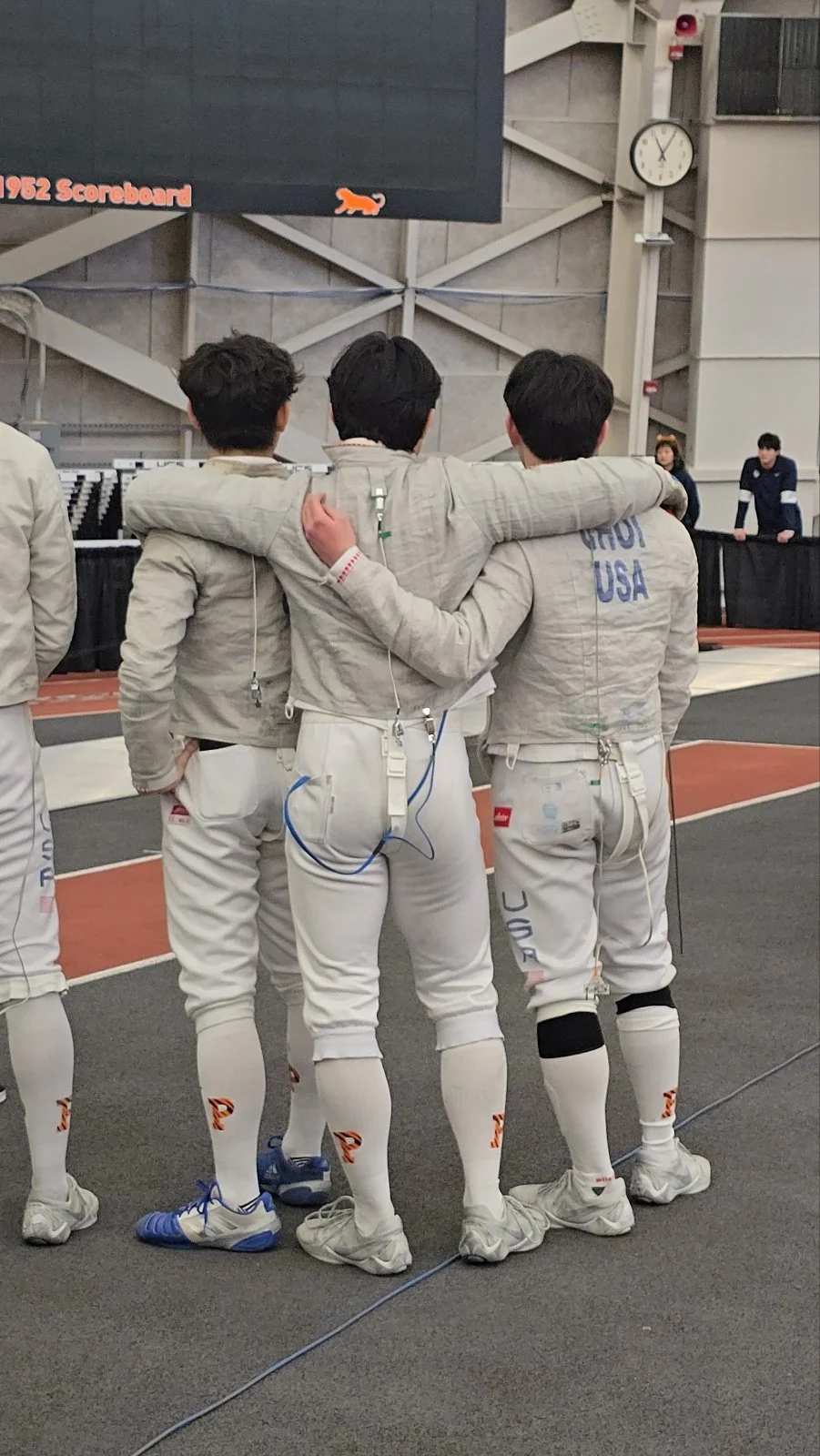 Three fencers in matching white uniforms with the USA emblem, standing with their arms around each other at an indoor fencing venue, with a scoreboard and clock in the background.