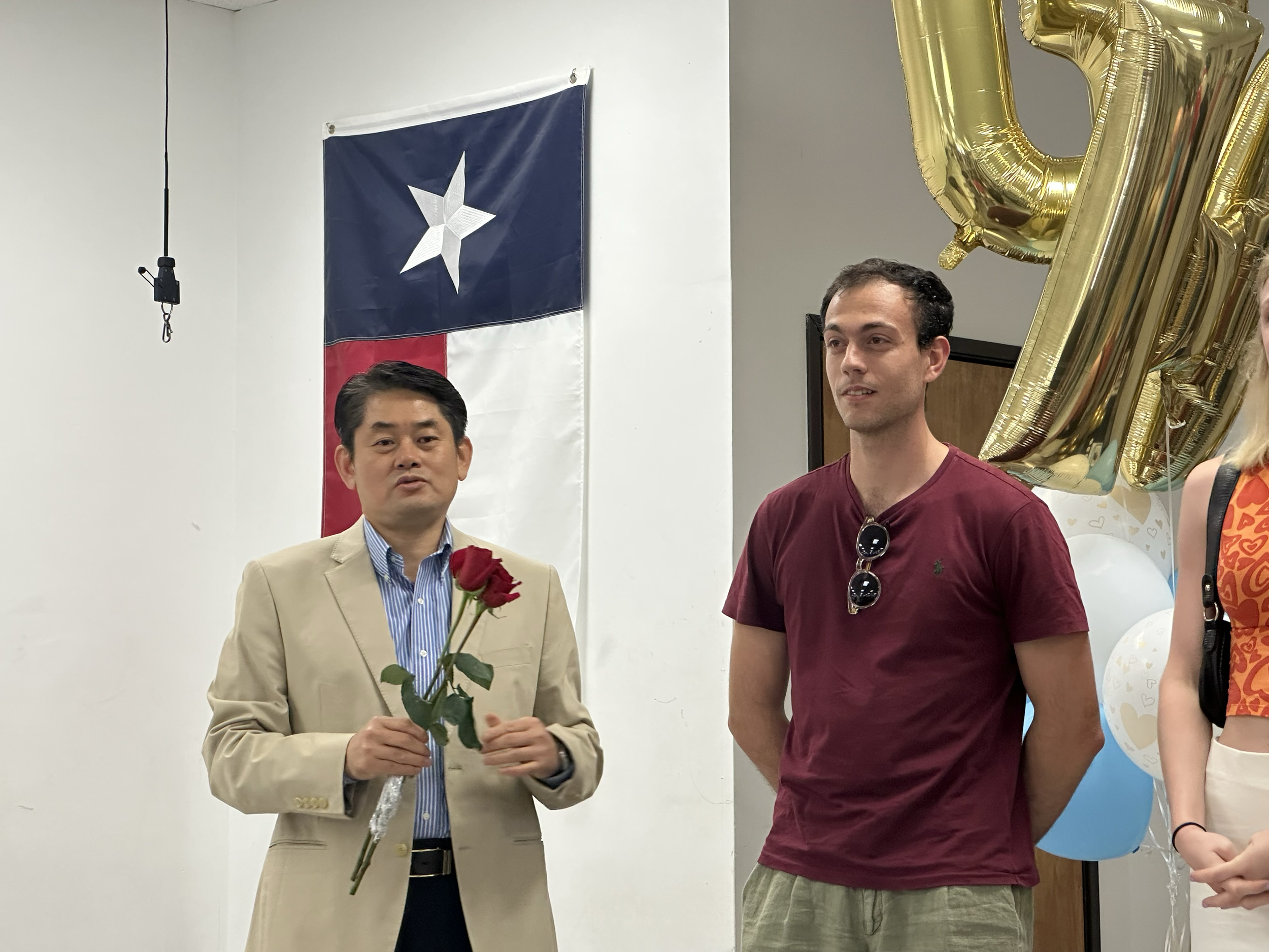 A man in a beige suit and blue striped shirt holding two red roses stands beside another man in a maroon t-shirt with sunglasses hanging from his collar. Behind them are balloon decorations and a Texas flag.