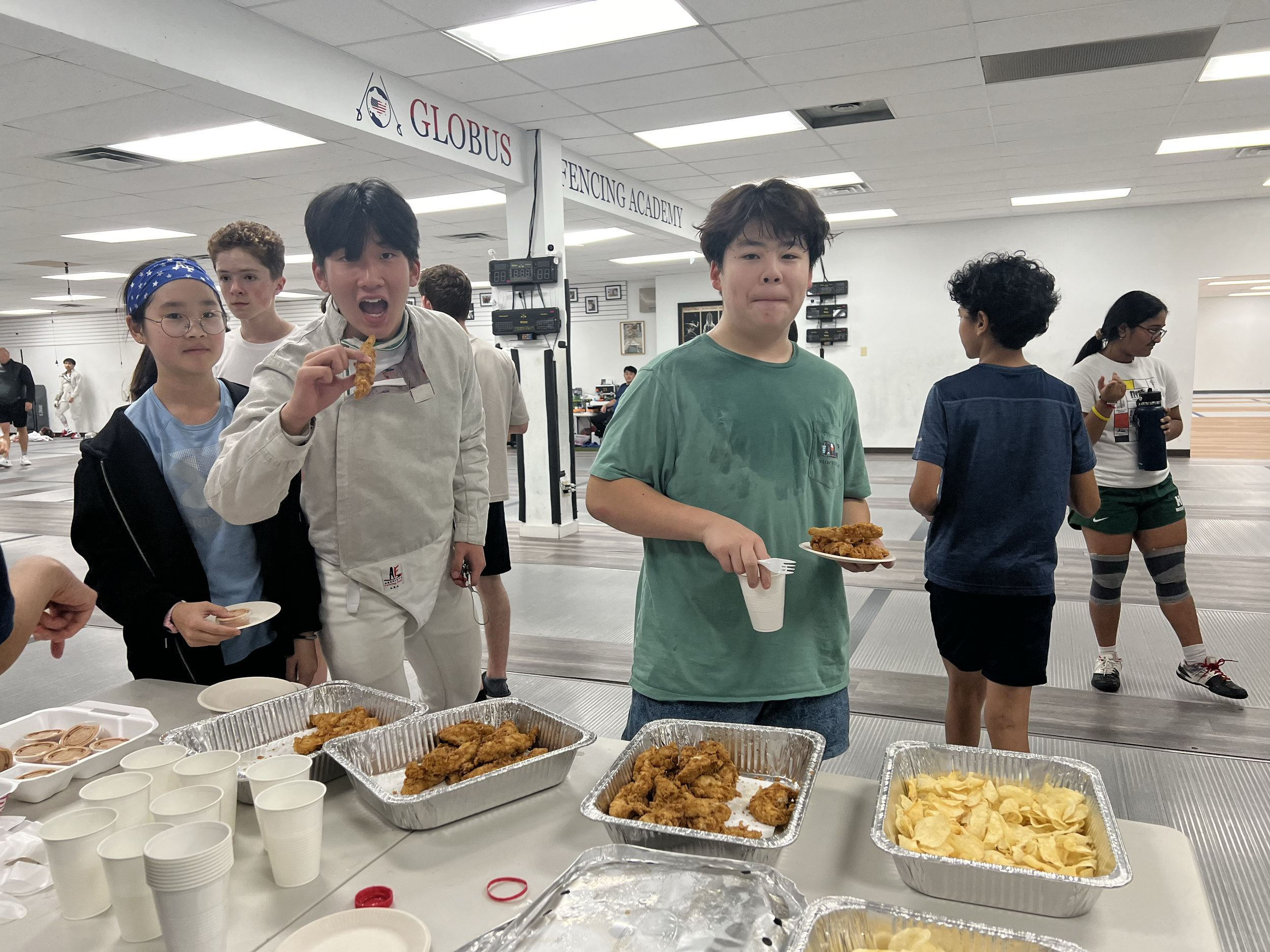 Group of children at a fencing academy eating fried chicken and chips from a buffet table.