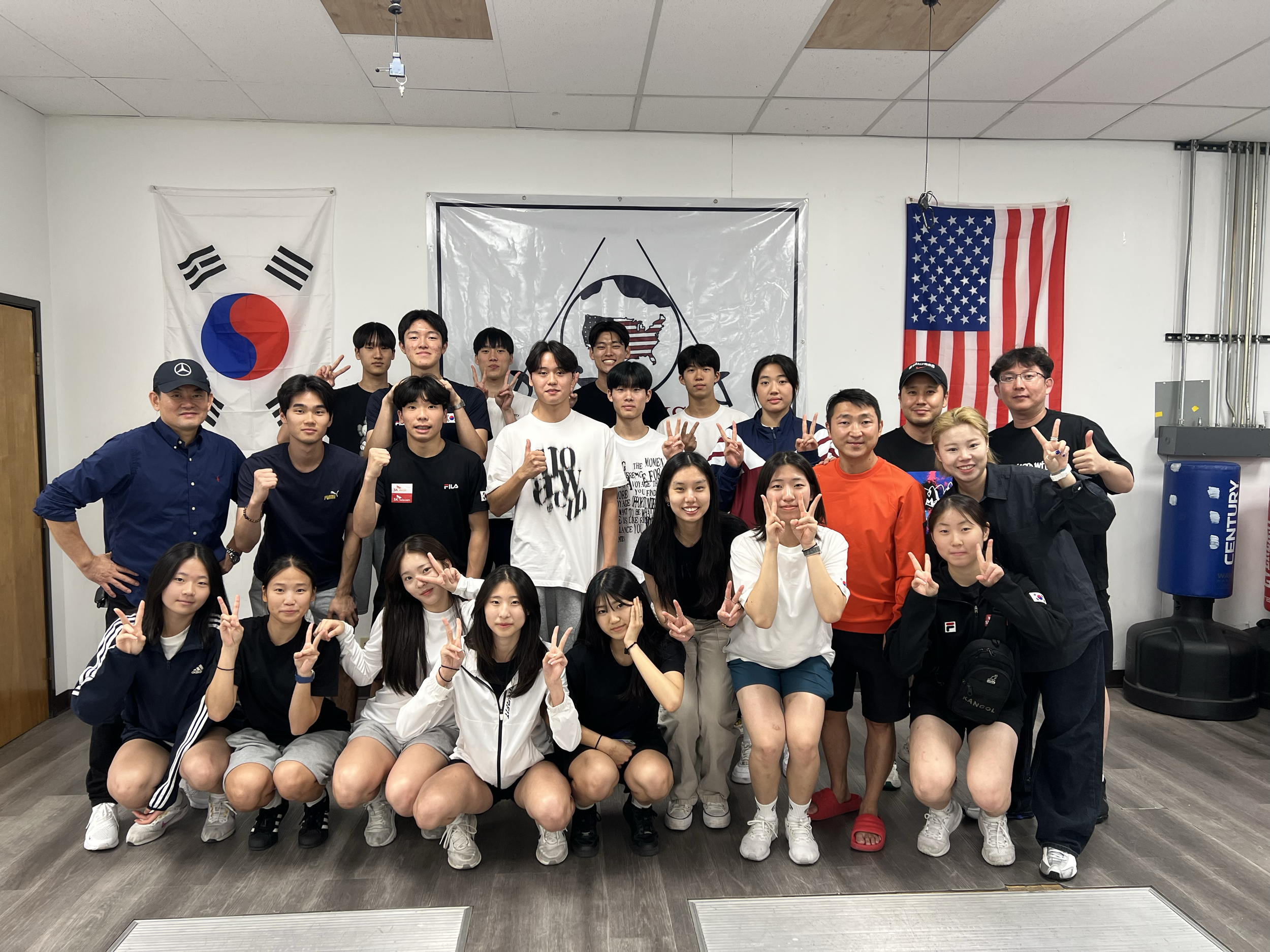A group of young people and adults posing together indoors in front of a South Korean flag, a black and white flag with a shield and two swords, and an American flag, with some participants making peace signs.