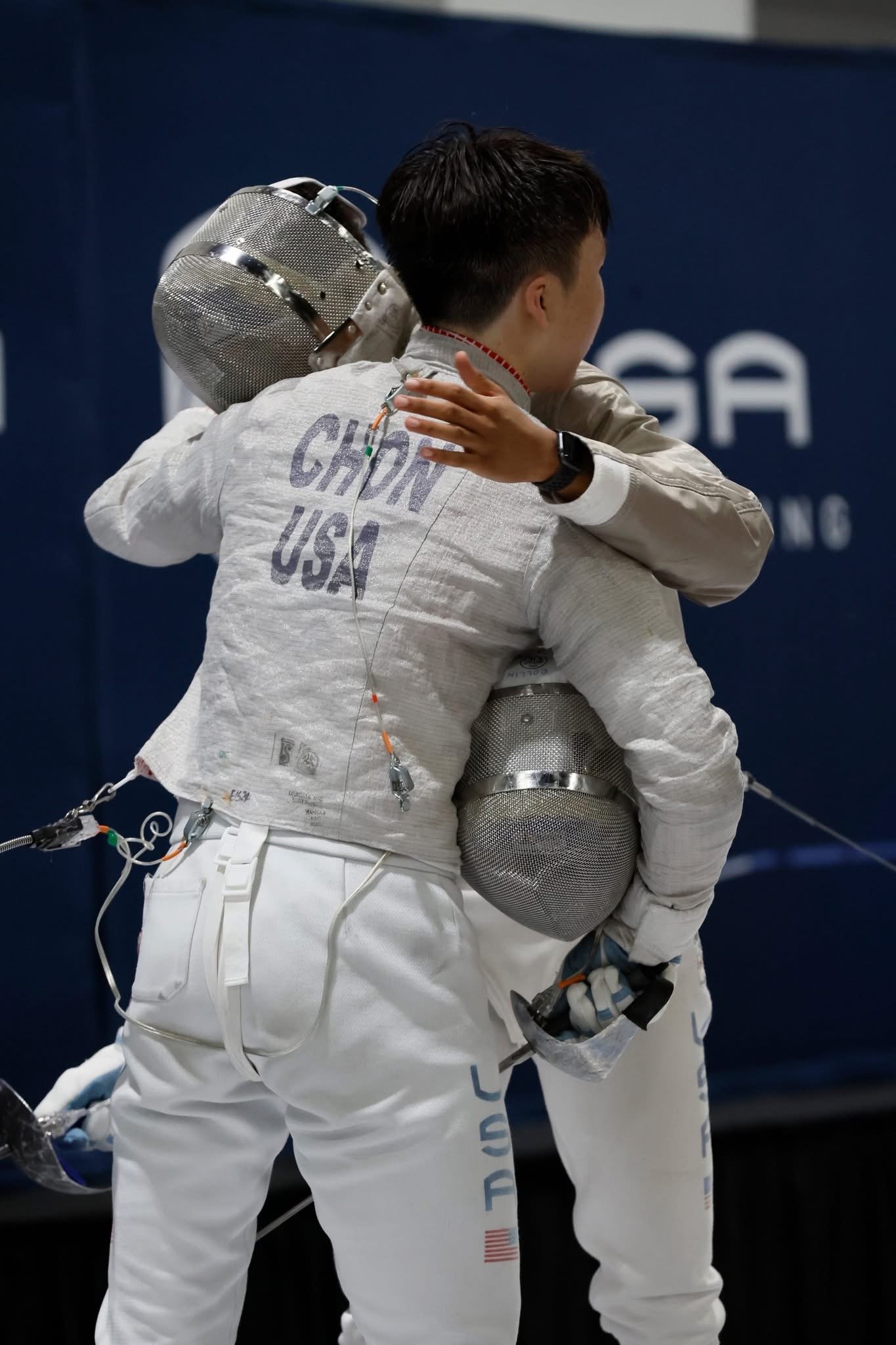 Two Olympic fencers embrace after a match, wearing fencing gear with 'CHOIN USA' on the back of one.