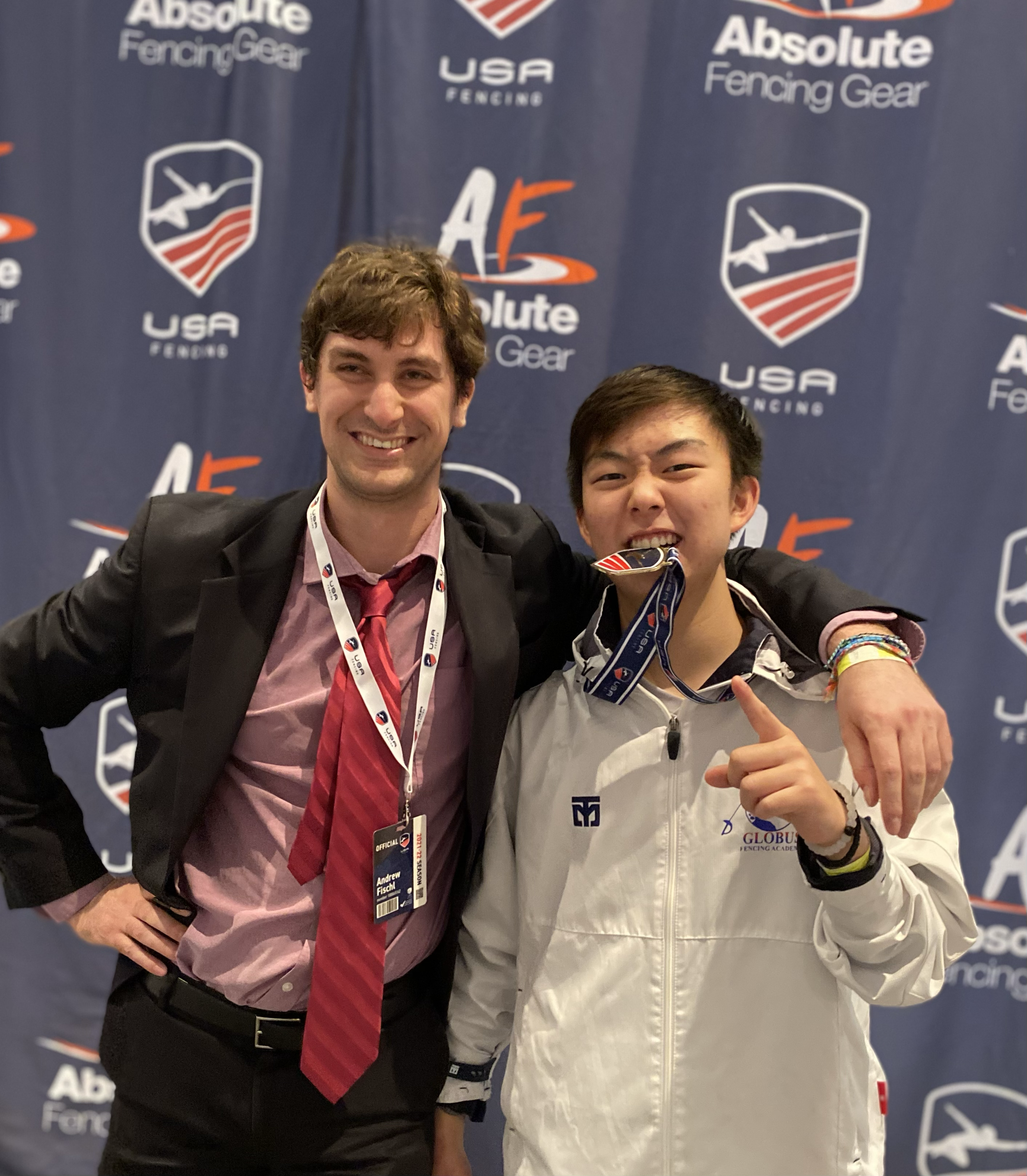 Two men smiling, one in a suit and red tie, the other in a white jacket with medals around his neck, standing in front of a blue backdrop with logos for USA Fencing and Absolute Fencing Gear.