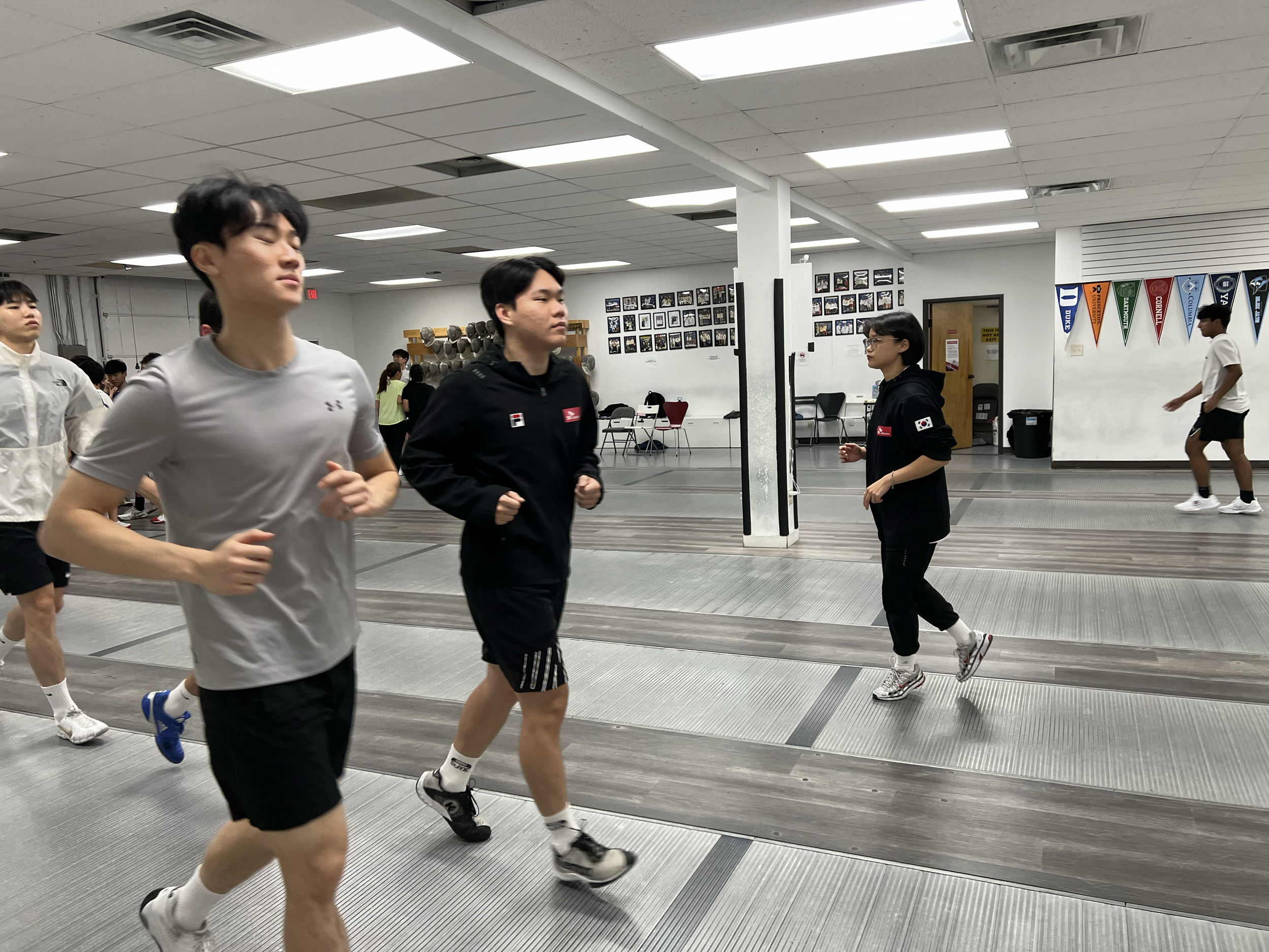 People running or jogging in indoor fencing training area with flags and photos on white walls.
