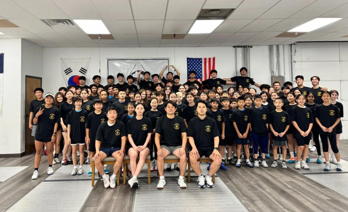 Group photo of a large athletic team with coaches, all wearing black shirts with a yellow logo, in a gym with South Korean and American flags in the background.