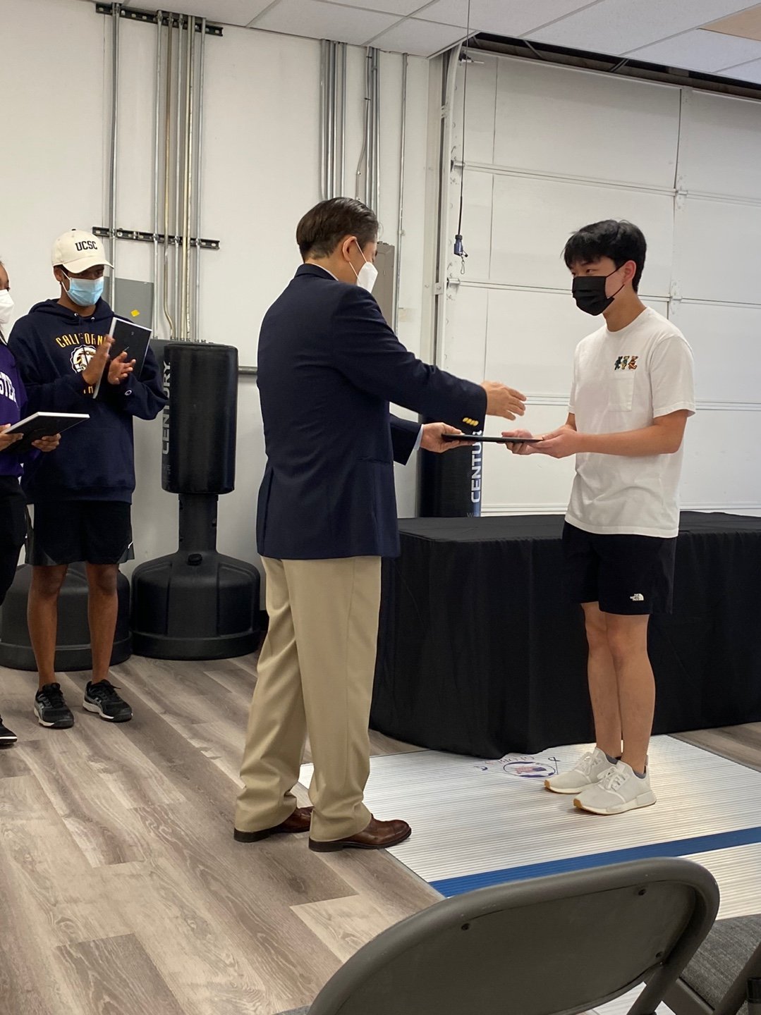 A young man in a white T-shirt and black shorts receives an award or certificate from an older man in a suit during a formal event. Two other young men in casual sportswear and masks observe, with one holding a notebook and the other a tablet.