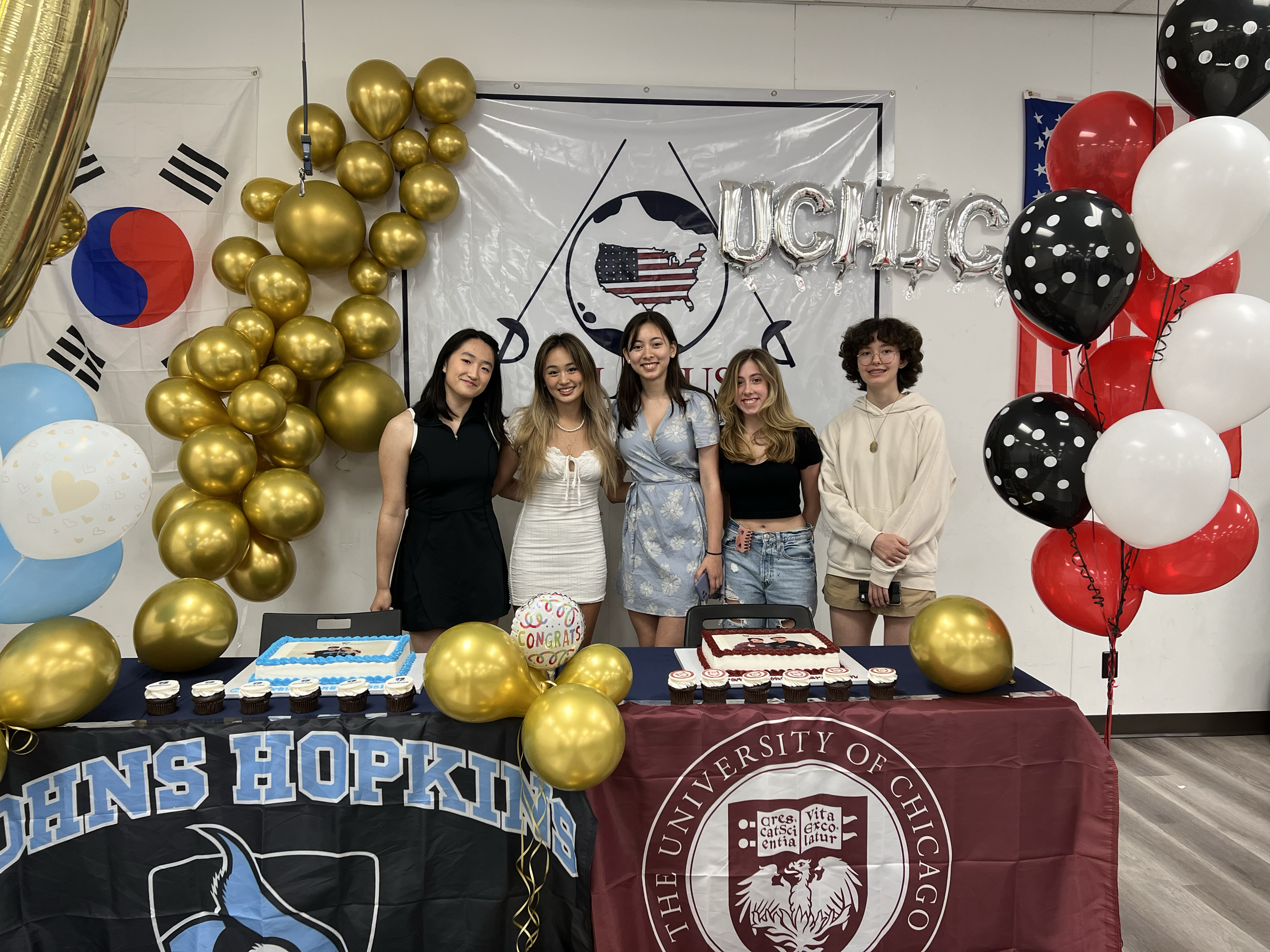 Five young women standing behind a table at a celebration event, with balloons, banners, and cupcakes. The table has a John Hopkins banner and a University of Chicago banner, with decorated cakes and balloons in black, red, white, blue, and gold. The
