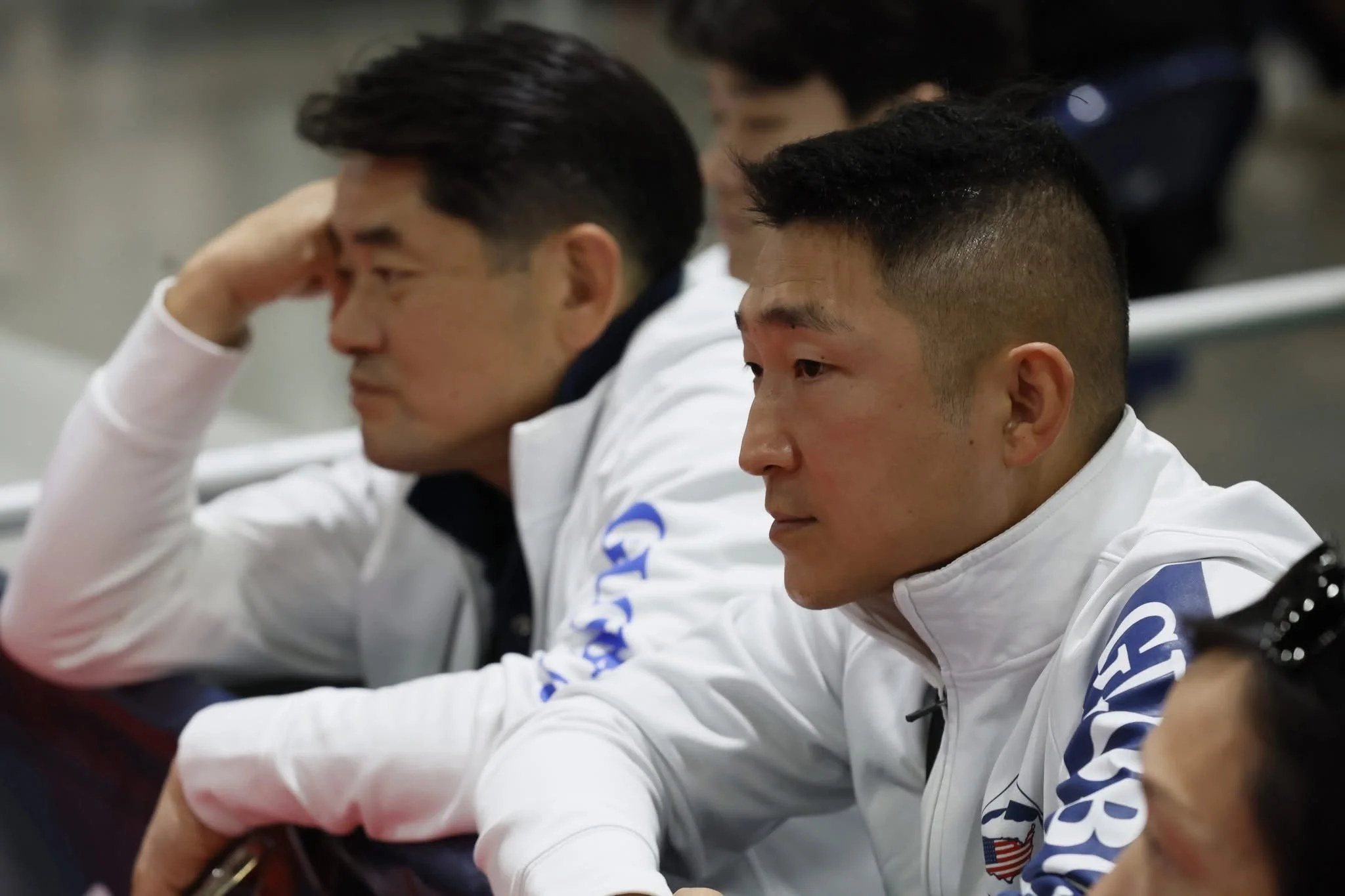 Two male athletes in white jackets with blue logos sitting at a sports event, looking focused and contemplative.