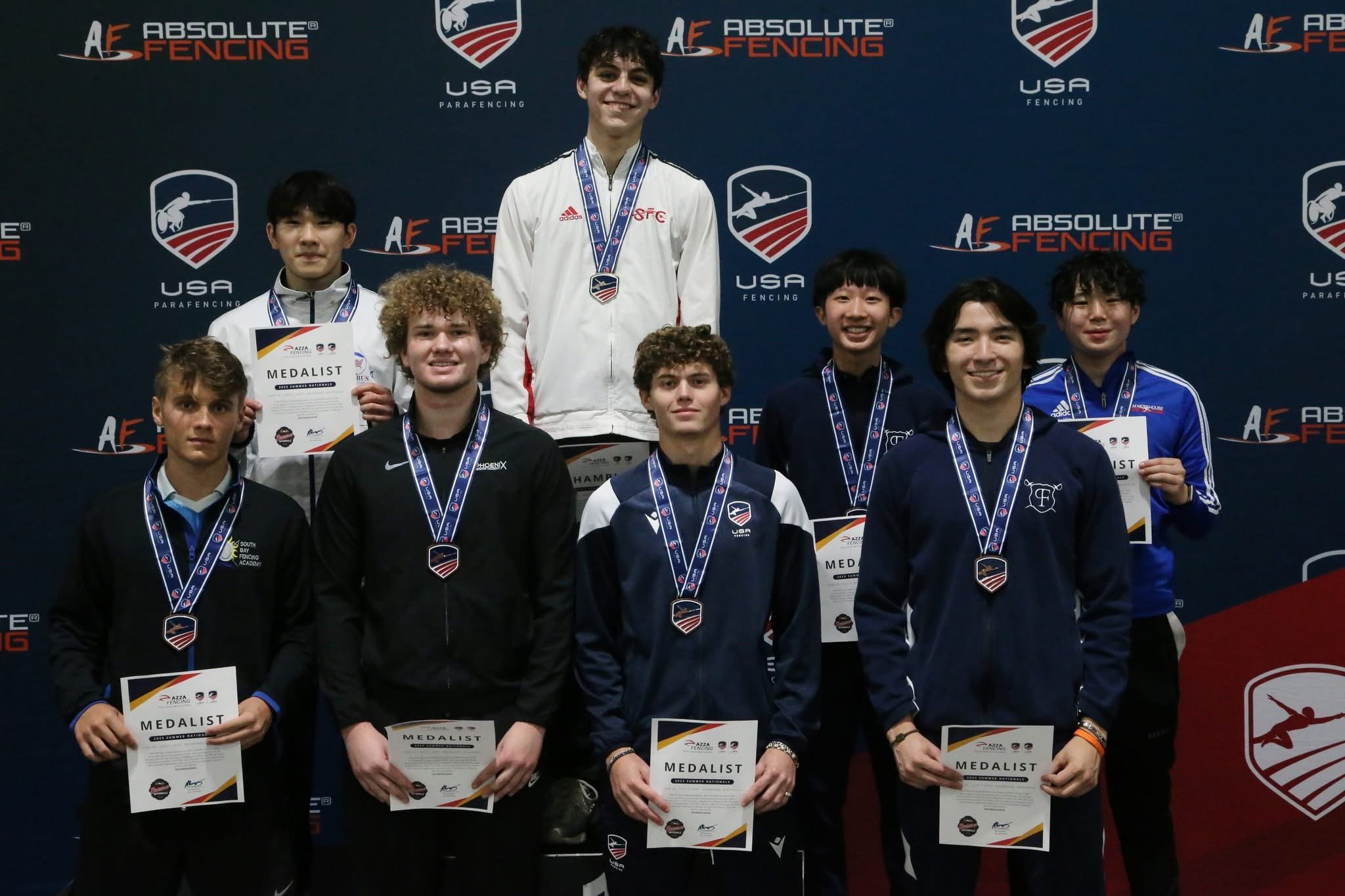 A group of nine young male athletes standing on a podium holding medals and certificates at a fencing competition. They are wearing sports jackets and track suits, smiling, with a dark blue backdrop displaying the logos for Absolute Fencing, USA Para