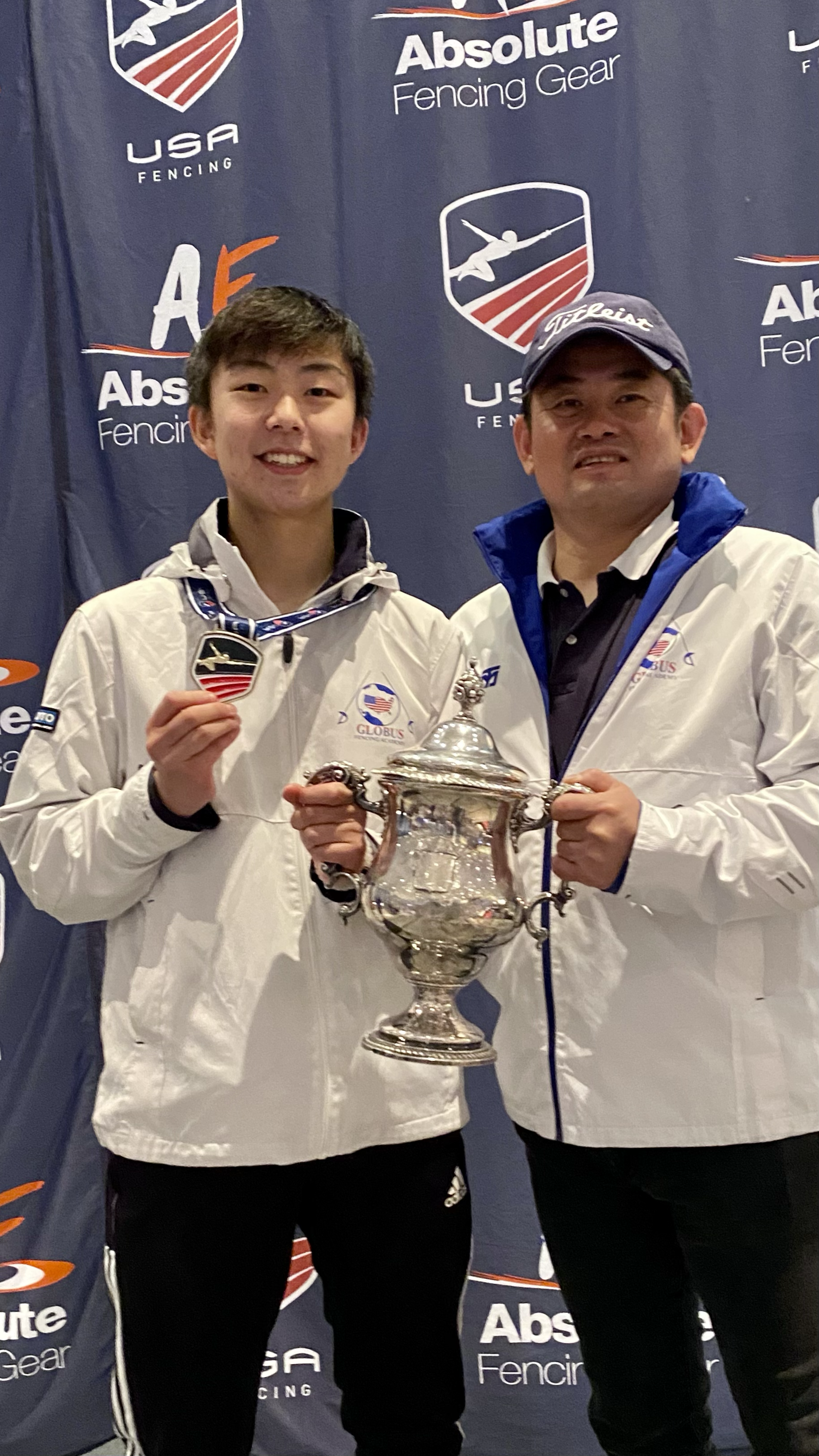 Two men in white jackets standing in front of a backdrop with logos, holding a large silver trophy and medals, celebrating a victory or award.