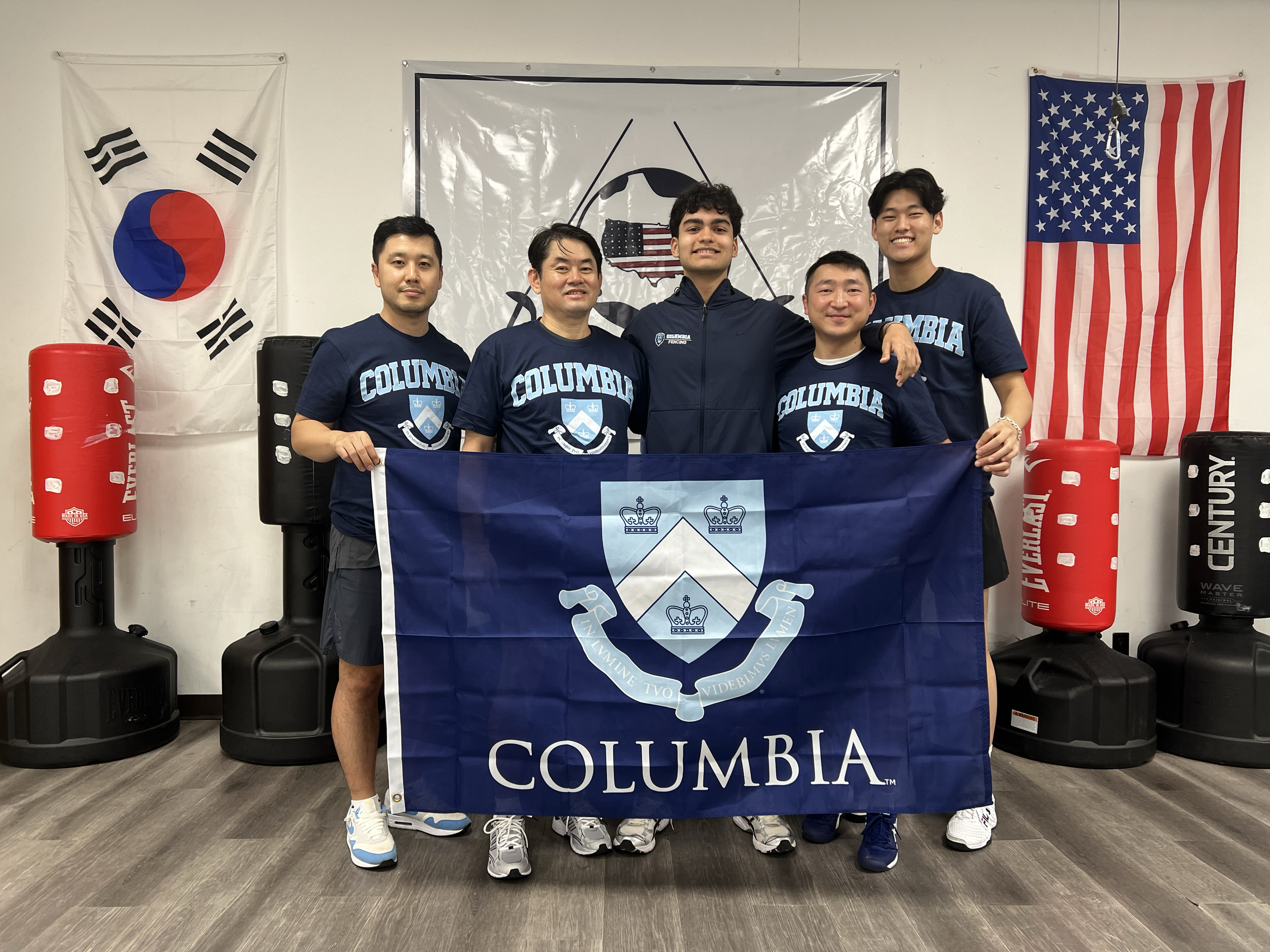 Group of five men standing indoors, holding a Columbia University flag, with two flags on the wall behind them—one of South Korea and one of the United States. They are wearing navy blue Columbia University shirts and casual athletic shoes, smiling f