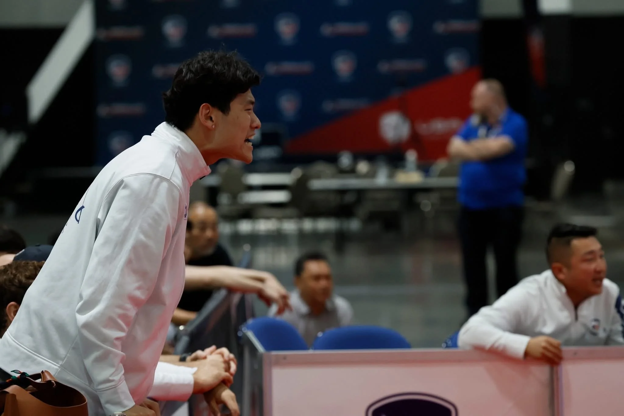 An Asian man in a white jacket is leaning forward and yelling or shouting, sitting at a table with others nearby. In the background, there is a blurred man standing with arms crossed, and another person sitting behind a barrier, all in a sports or in