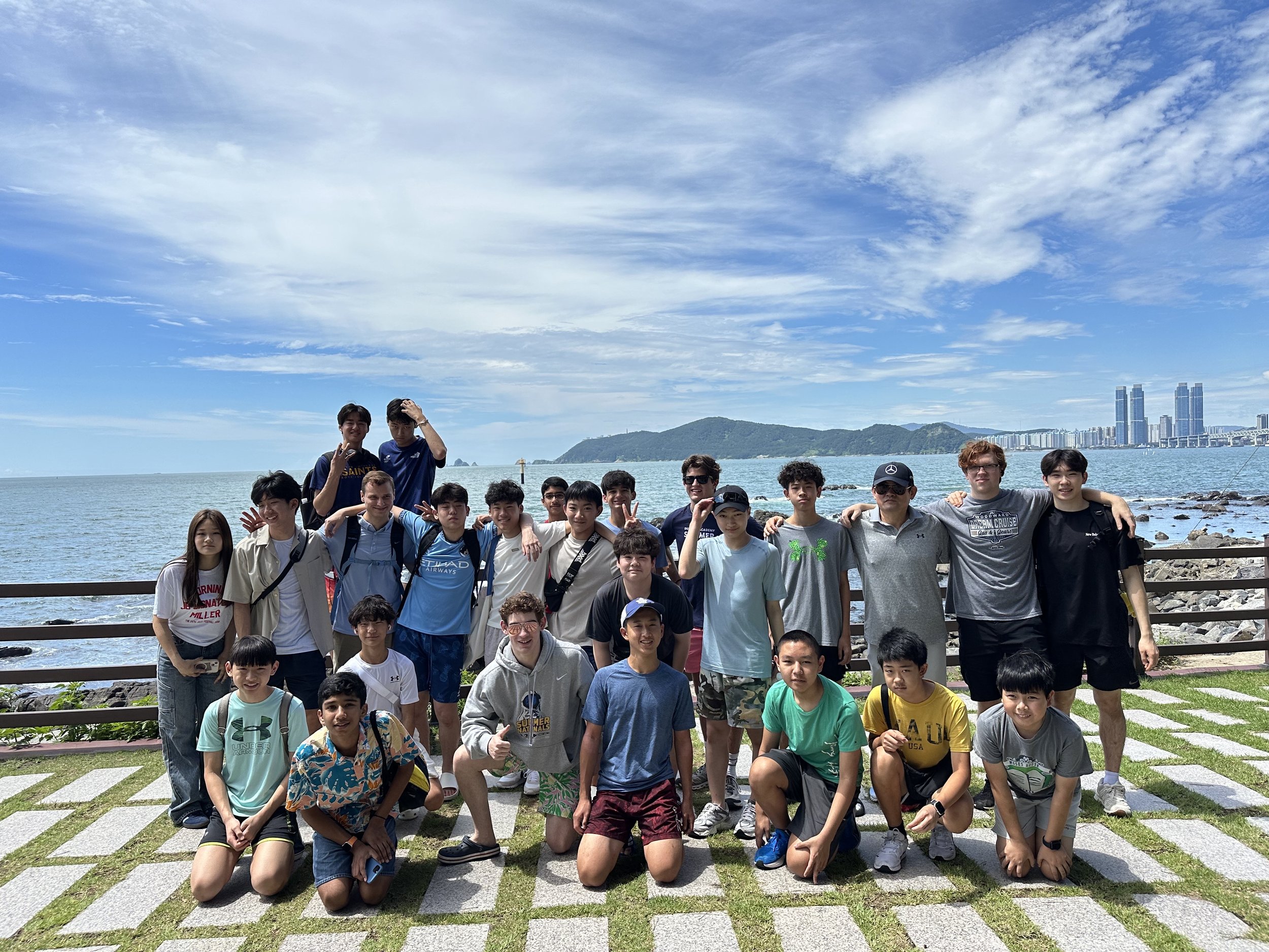 Group of children and adults posing for a photo on a seaside promenade with a city skyline and mountains in the background.