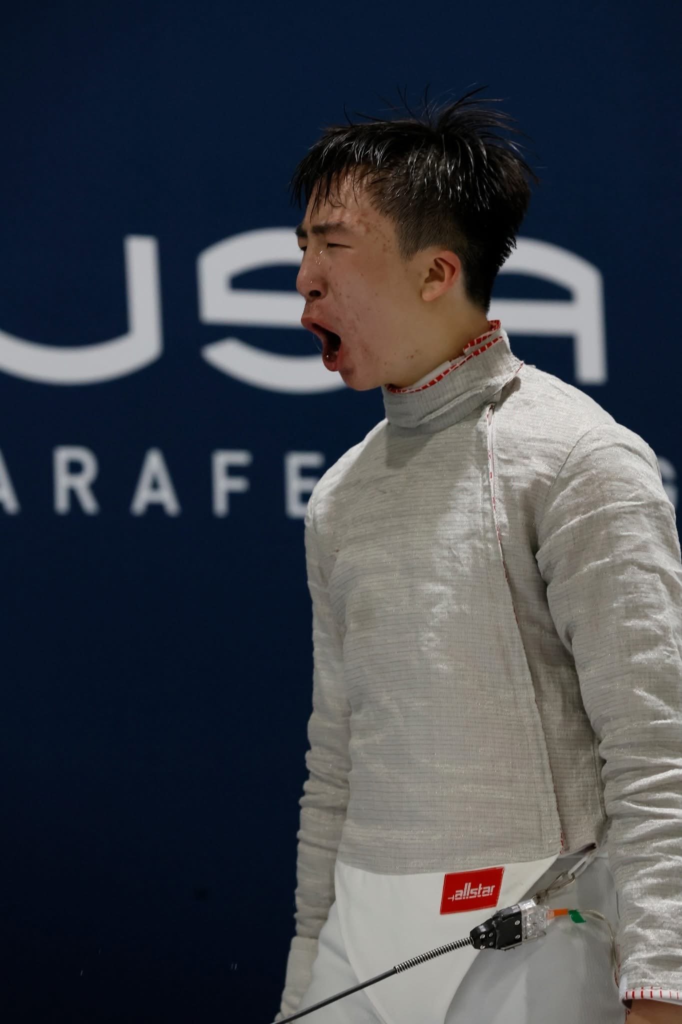 Male athlete in fencing suit yawning or shouting at a fencing event.