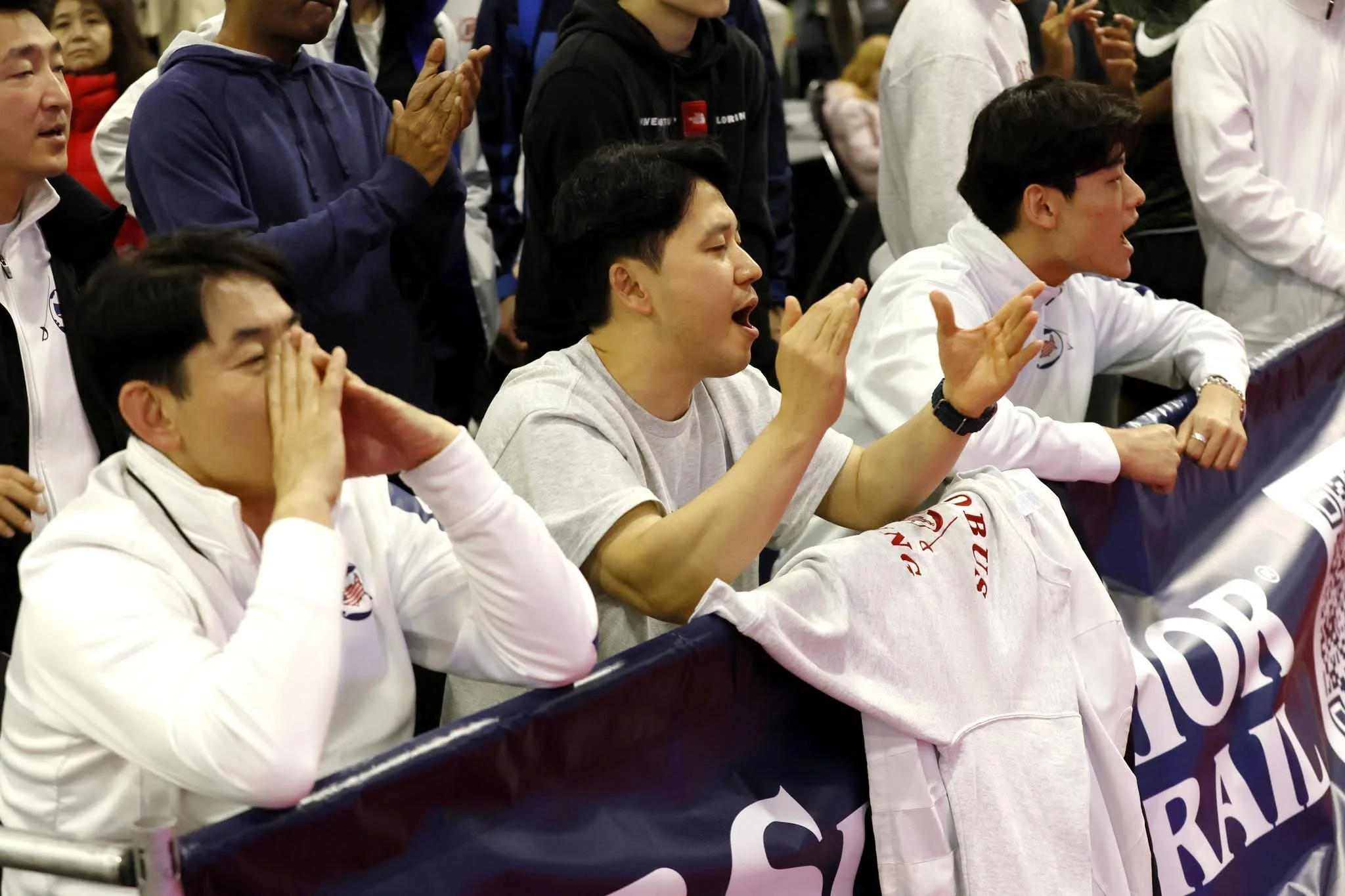 Group of people, including athletes wearing white jackets, watching a sports event, with some cheering and one using a megaphone.