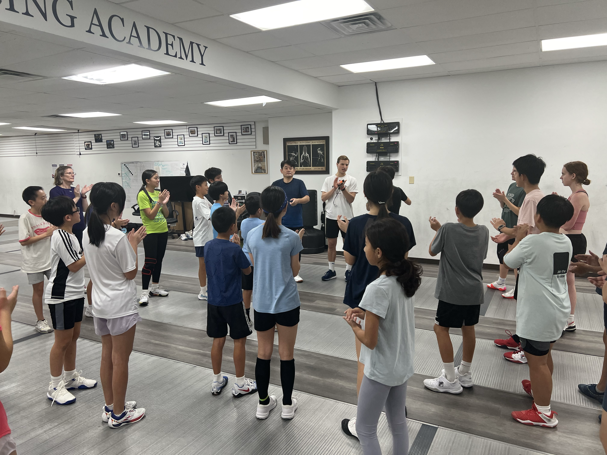 Children and adults participating in a dance class at a dance academy, with some clapping and others practicing moves, inside a studio with gray floors and a white wall.