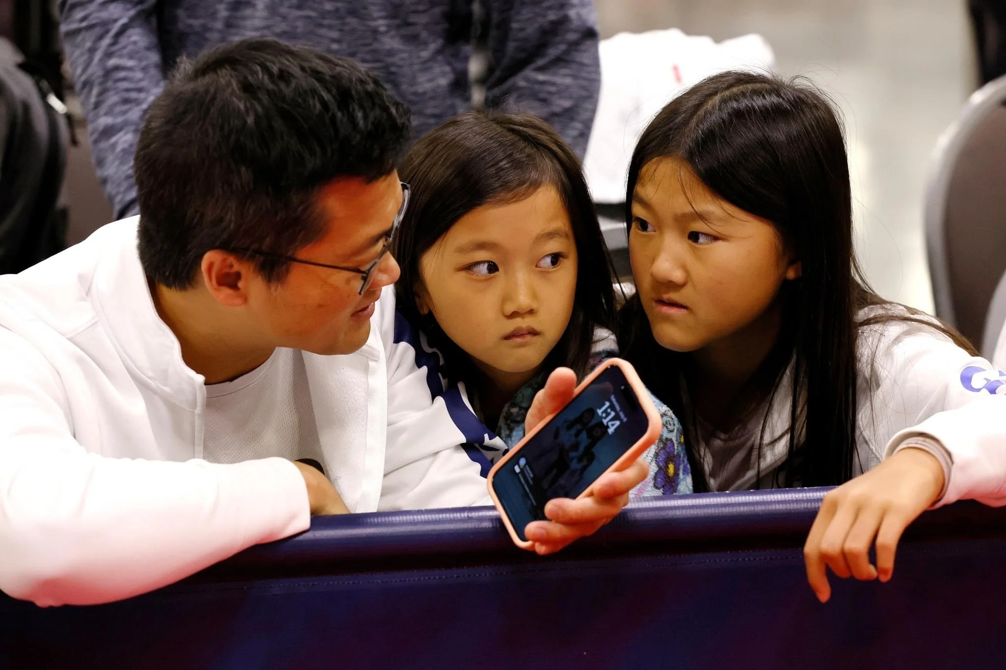 An adult male and two young girls are sitting close together, looking at a smartphone that the man is holding. They appear to be indoors at an event or gathering.
