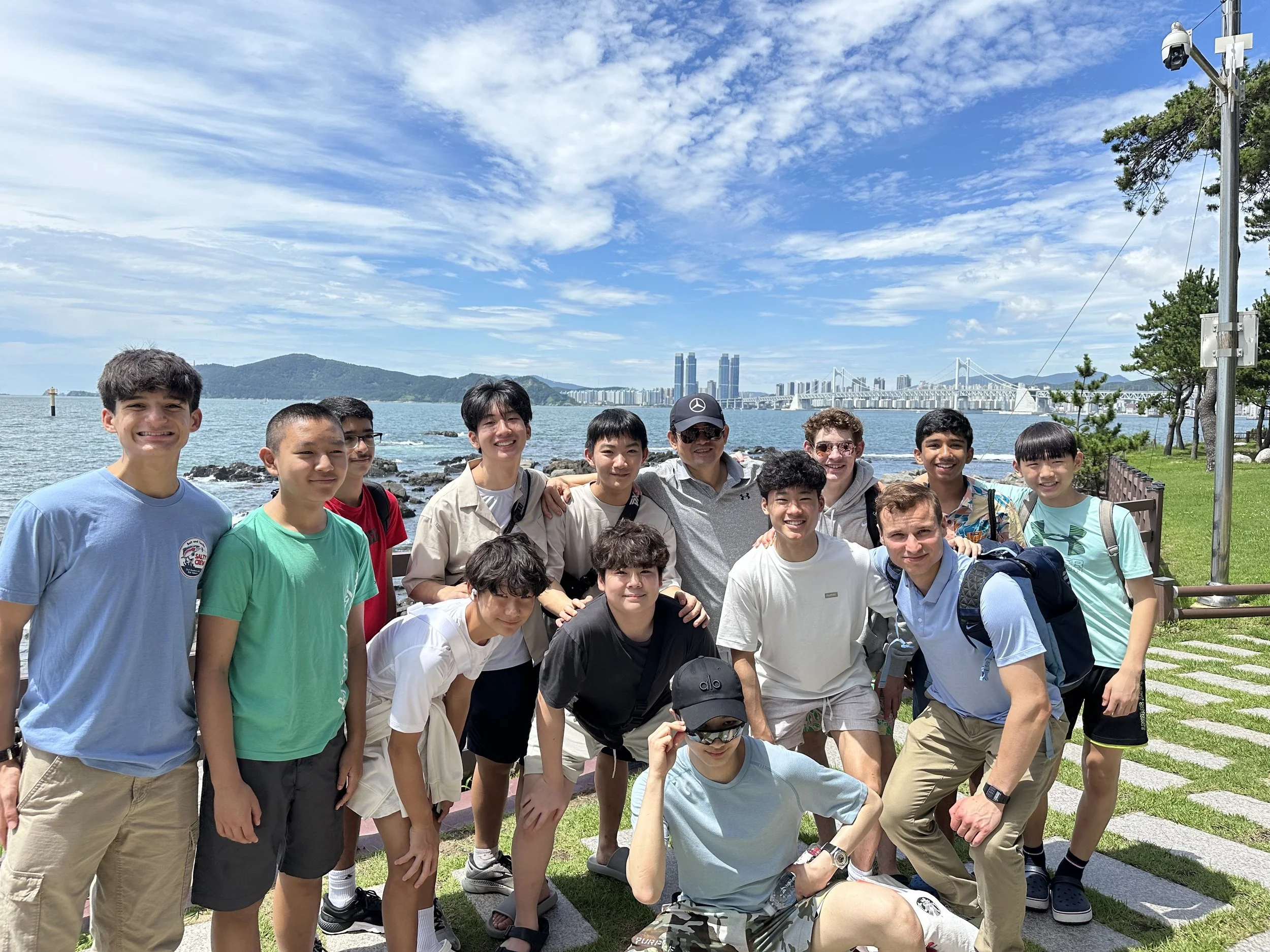 Group of teenagers and adults posing by the water with a city skyline and bridge in the background on a sunny day.