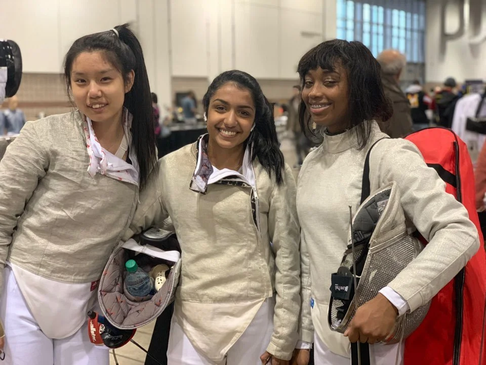 Three women wearing beige jackets and white pants standing together at a crowded indoor event, smiling at the camera.