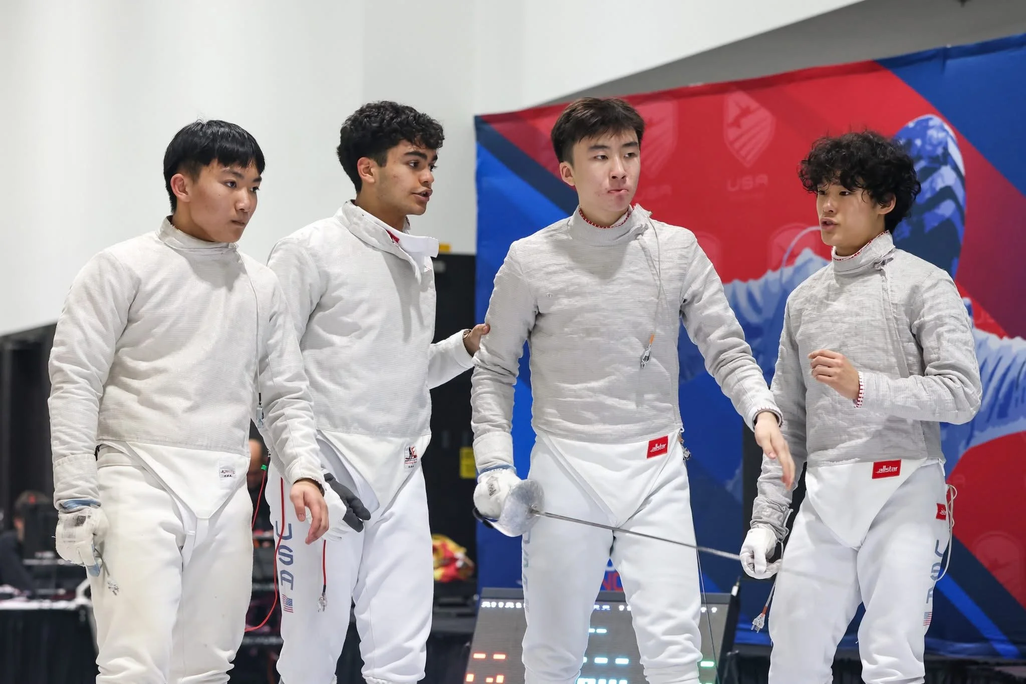 Four male fencers in white uniforms standing together, one holding a fencing sword, in an indoor sports venue with a colorful backdrop.