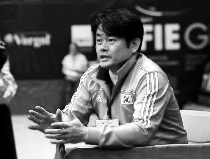 A man with dark hair, wearing a South Korea sports jacket, is sitting at a table and speaking, with his hands gesturing as he talks.
