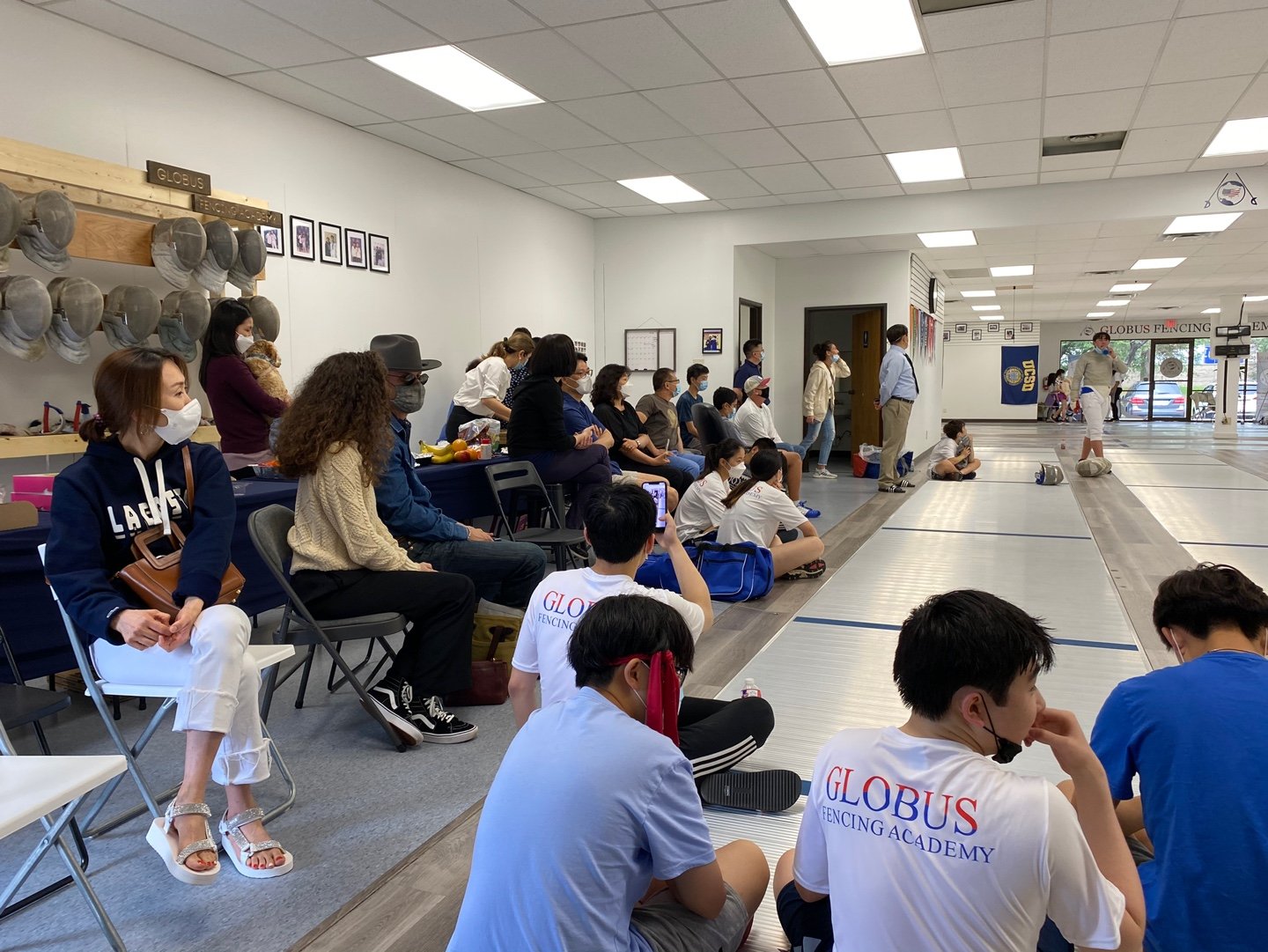 People gathered in a fencing academy watching a fencing match, some seated and others standing, with fencing equipment and banners visible in the background.