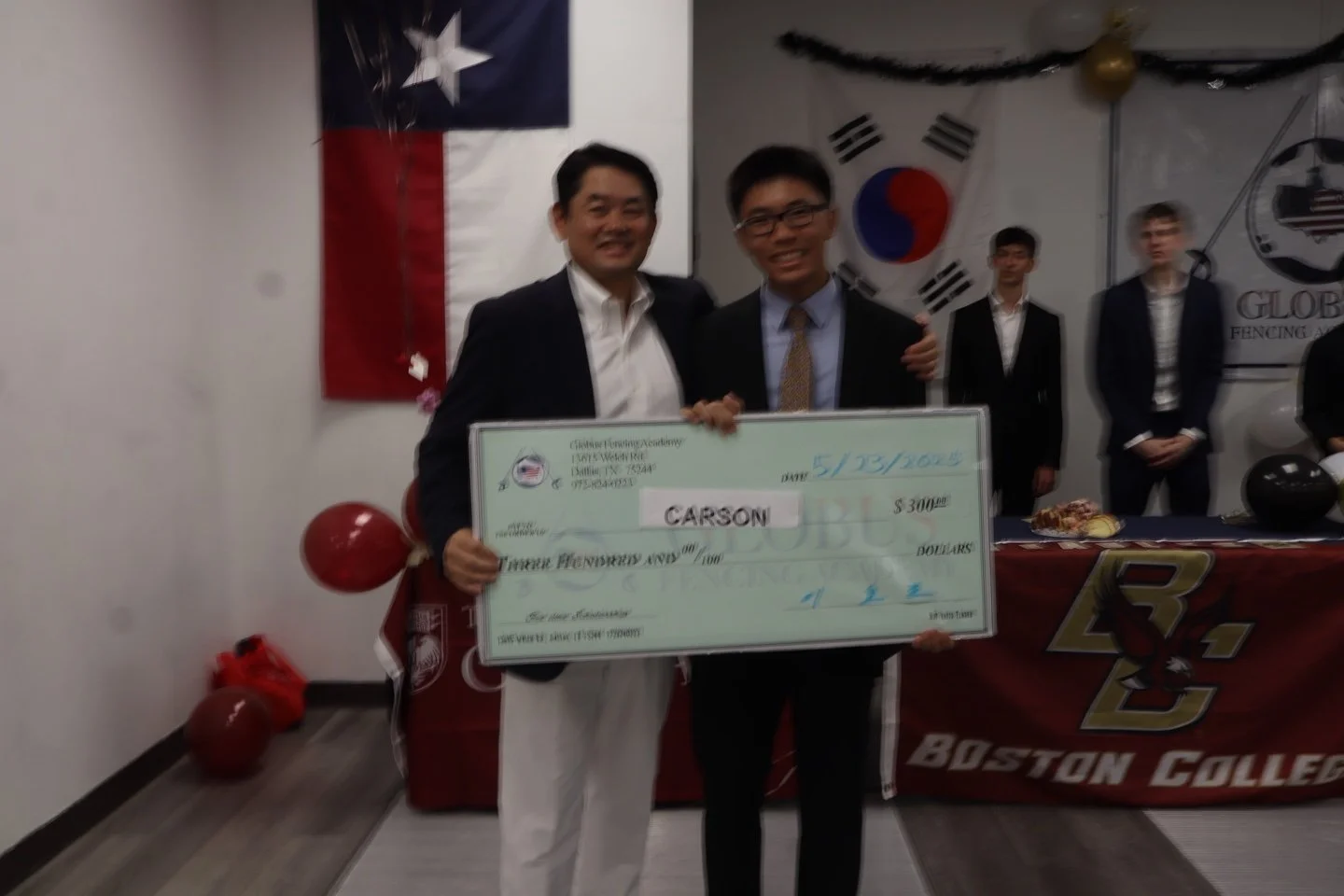 Two men in suits holding an oversized check for $300 with the name Carson, celebrating at an event with Texas and South Korea flags in the background.