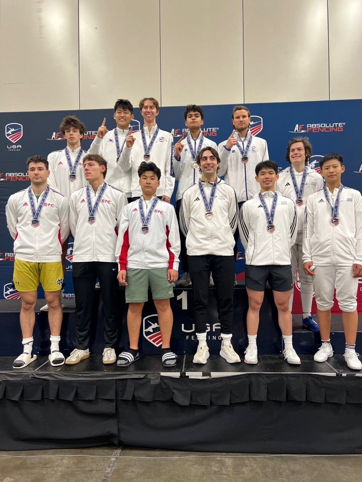 Young male fencing athletes and coach on podium wearing medals at USA Fencing competition.