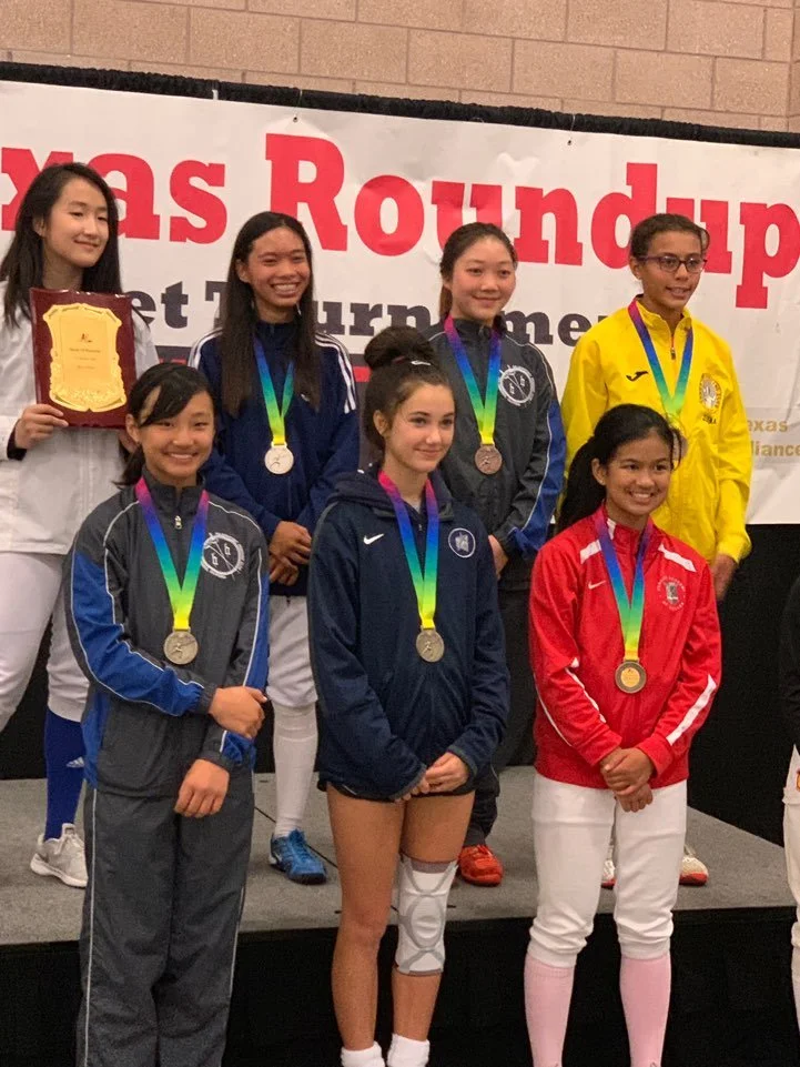 Young female athletes on a podium receiving medals at a football tournament, with a banner in the background reading 'Texas Roundup'.