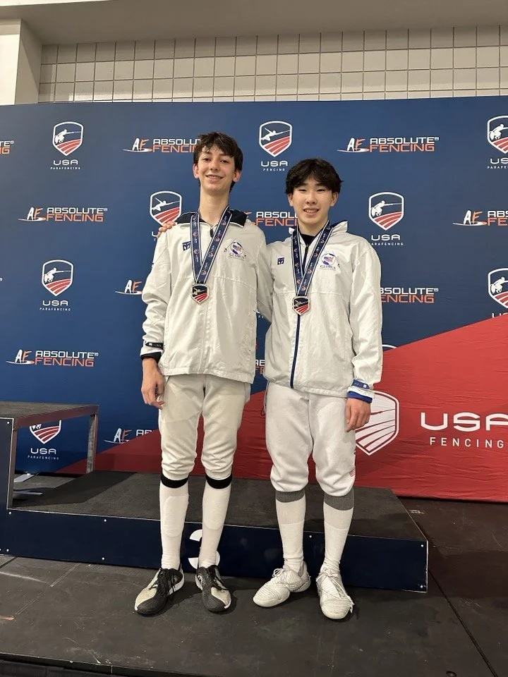 Two young male fencers wearing white fencing uniforms standing on a winners' podium, each with a medal around their necks, at an USA Fencing event.