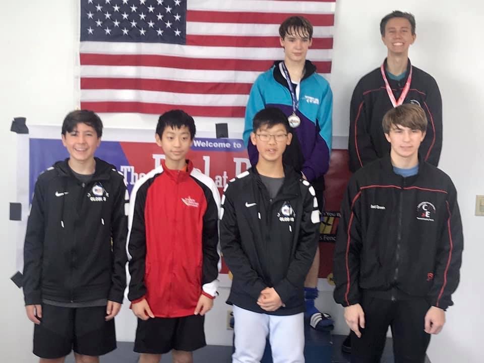 Six young male swimmers standing in front of an American flag and a banner at a swimming competition, some wearing medals and team jackets.