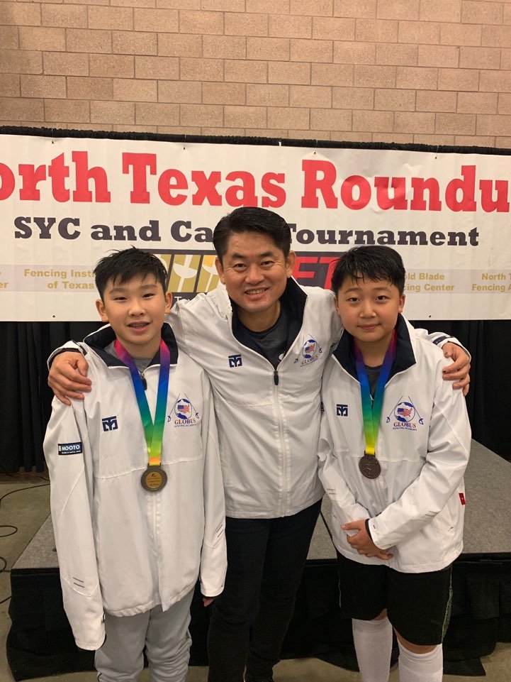 Three boys and an adult male at a fencing tournament, with everyone wearing medals and team jackets, standing in front of a banner that reads 'North Texas Roundup SYC and Cadet Tournament.'