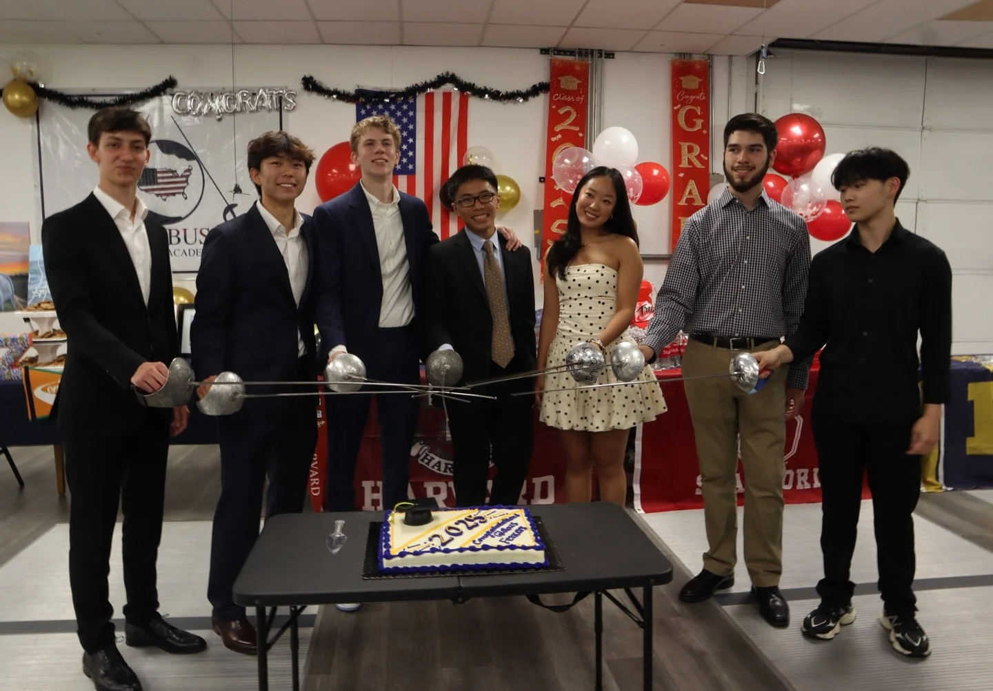 Group of eight young people standing behind a celebration cake on a table, with graduation decorations in the background.