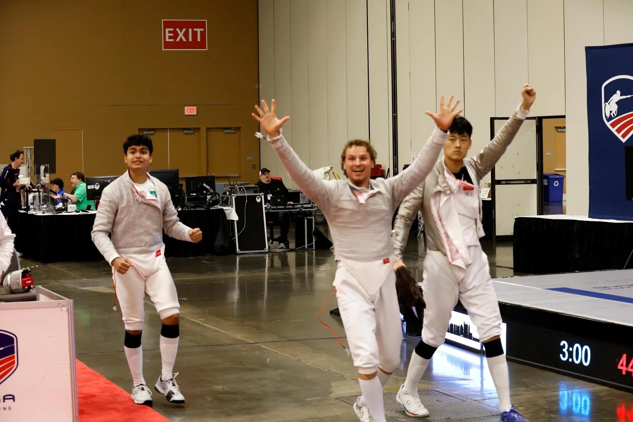 Three young men in fencing gear celebrating in an indoor sports arena, with others and equipment in the background.