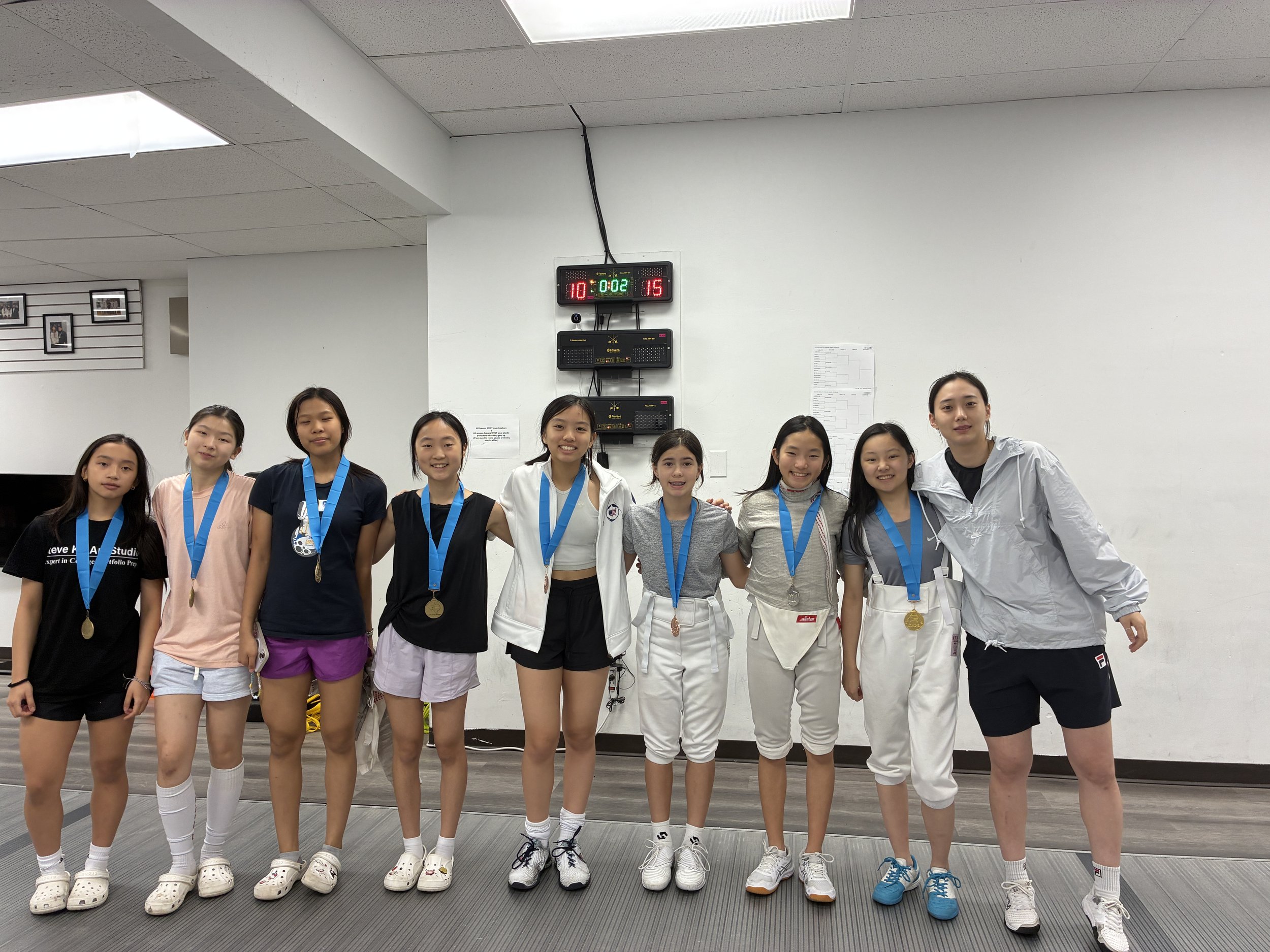 A group of young female athletes pose together after a fencing competition, wearing medals around their necks, in an indoor sports facility.