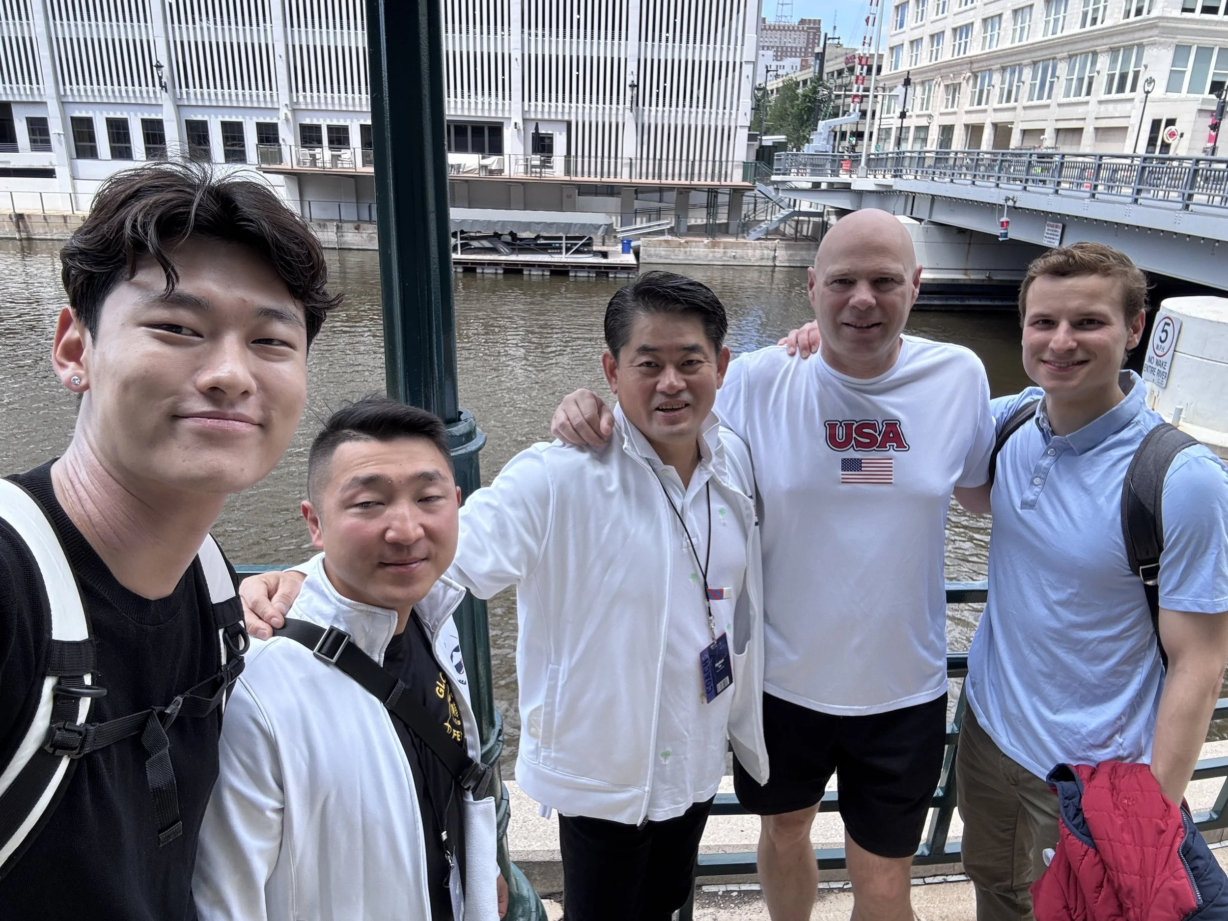Group of five men standing outdoors near a river with city buildings and a bridge in the background, smiling for a selfie.