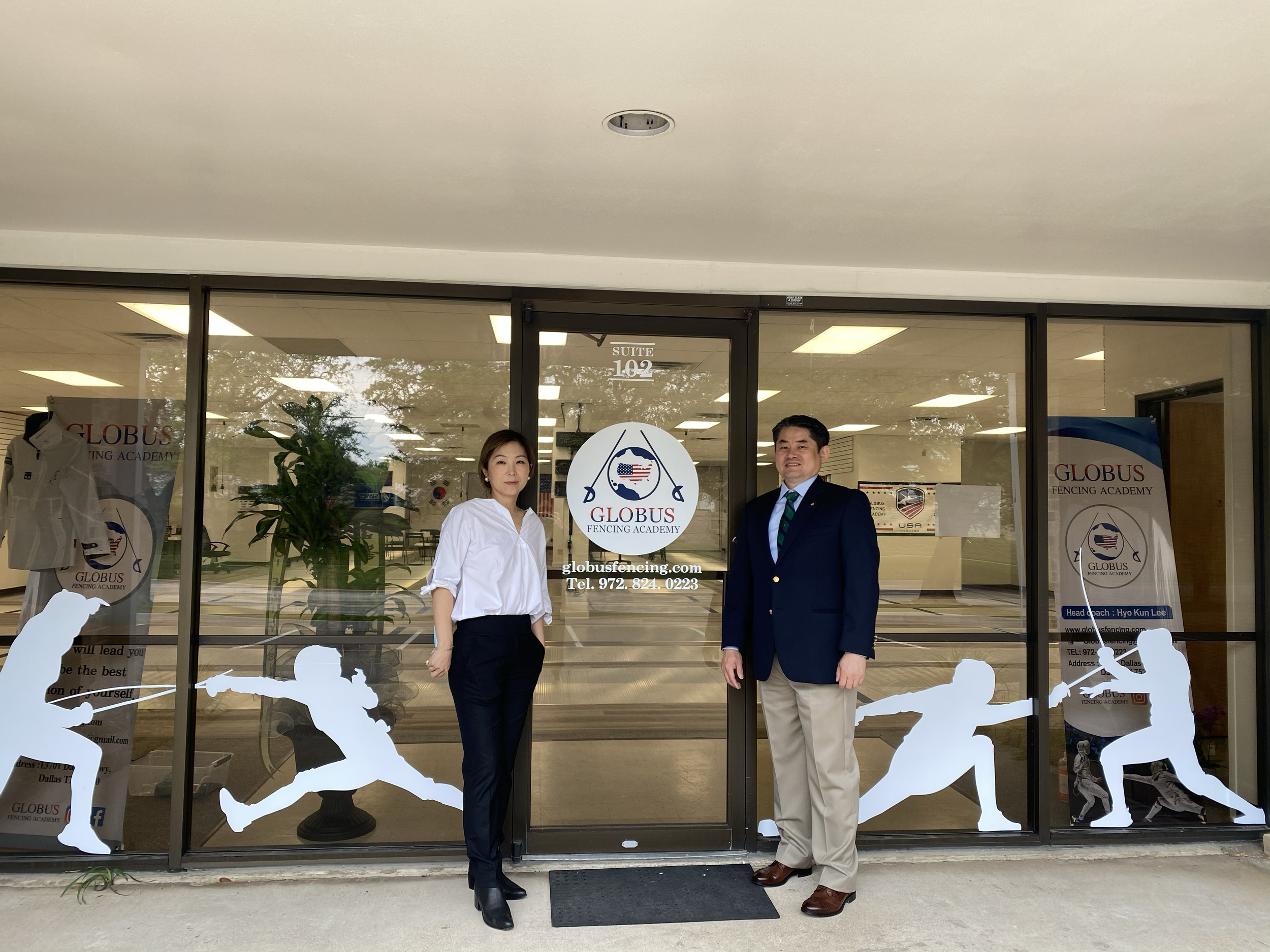 Two people standing outside the entrance of Globus Fencing Academy, a woman on the left in a white blouse and dark pants, a man on the right in a navy blazer with beige pants, both smiling. The glass door features the academy logo with fencing imager