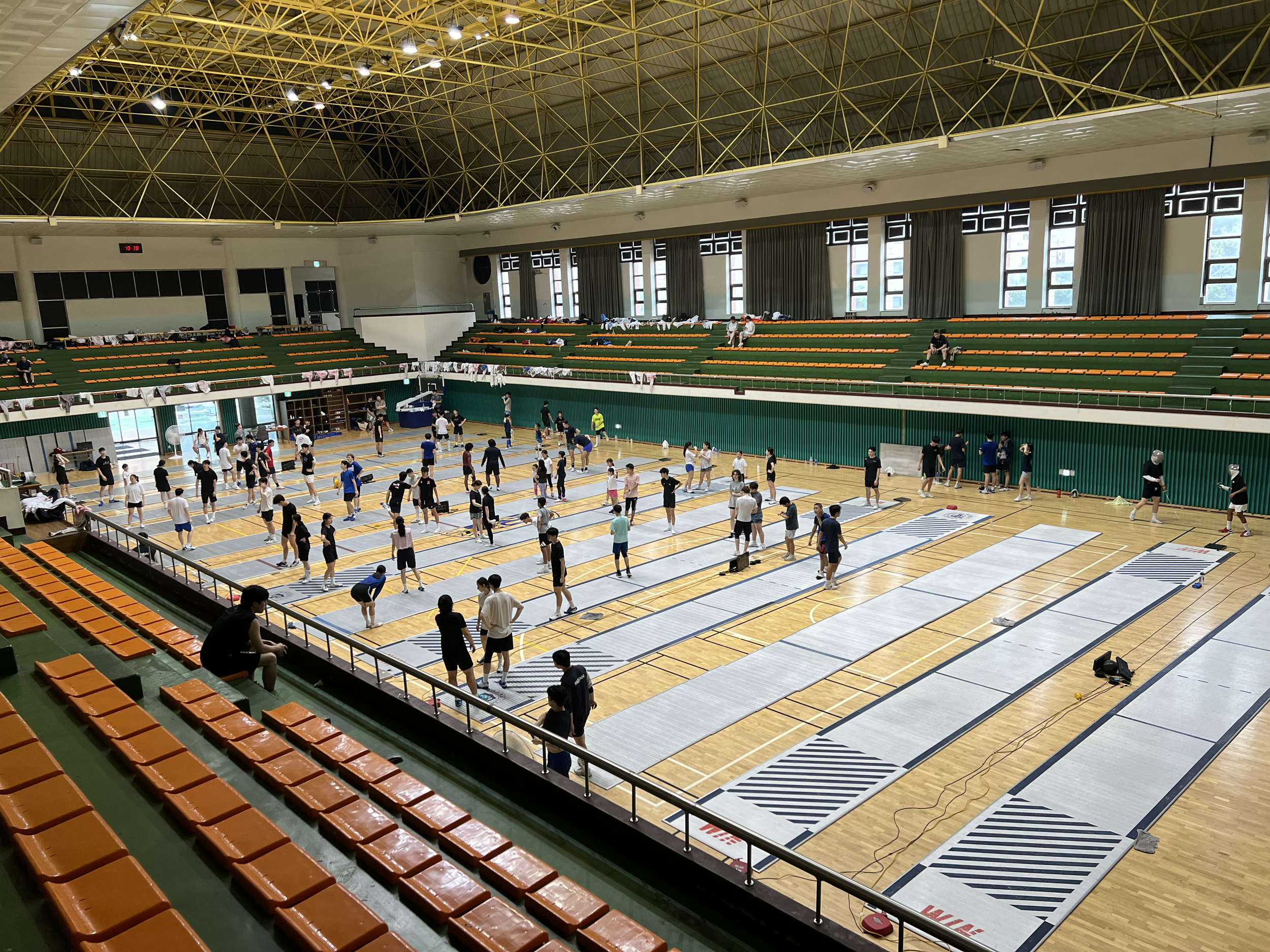 Indoor sports hall with several people practicing track and field events, with long mats laid out on the wooden floor for running drills or jumping exercises. Spectator seating is visible in the background and a few individuals are sitting in the ble