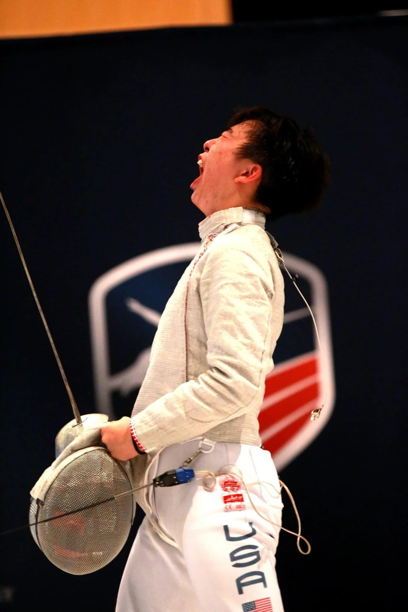 A male fencer in fencing gear, holding a fencing mask, with a background that features a fencing logo.