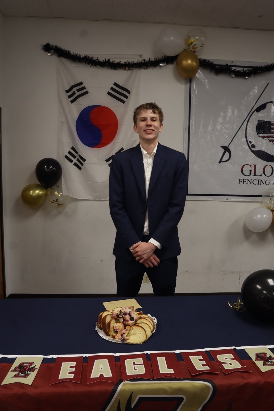 A young man in a navy suit and white shirt stands in front of a decorated wall with a South Korean flag, black, white, and gold balloons, and black tinsel. There is a table with sliced cake and a 'BEAGLES' banner in front of him.