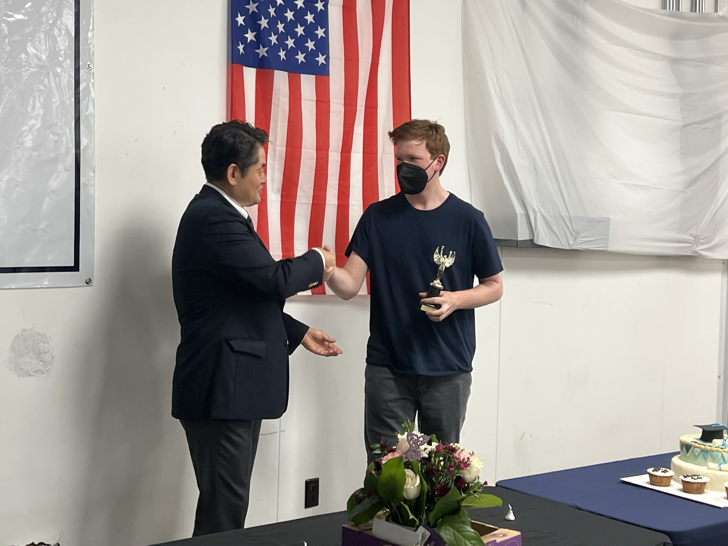 A young man in a navy blue t-shirt and a black face mask receives an award and shakes hands with an older man in a suit during a ceremony. The young man holds a small trophy, with a table of cupcakes and a bouquet of flowers nearby. An American flag 
