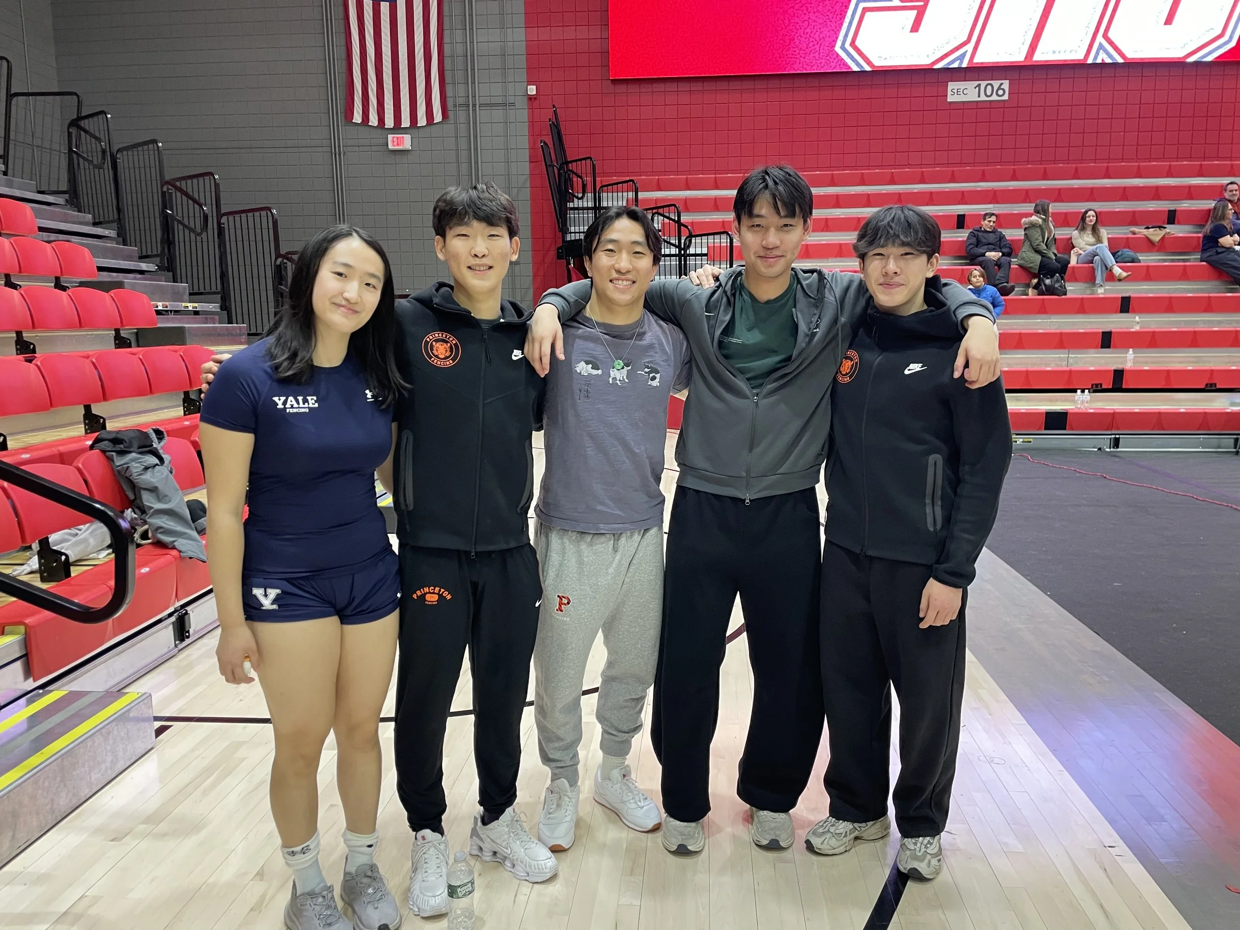 A group of five young athletes standing together in an indoor sports arena, with red bleachers and a large LED screen in the background. They are smiling and appear to be part of a track and field team.