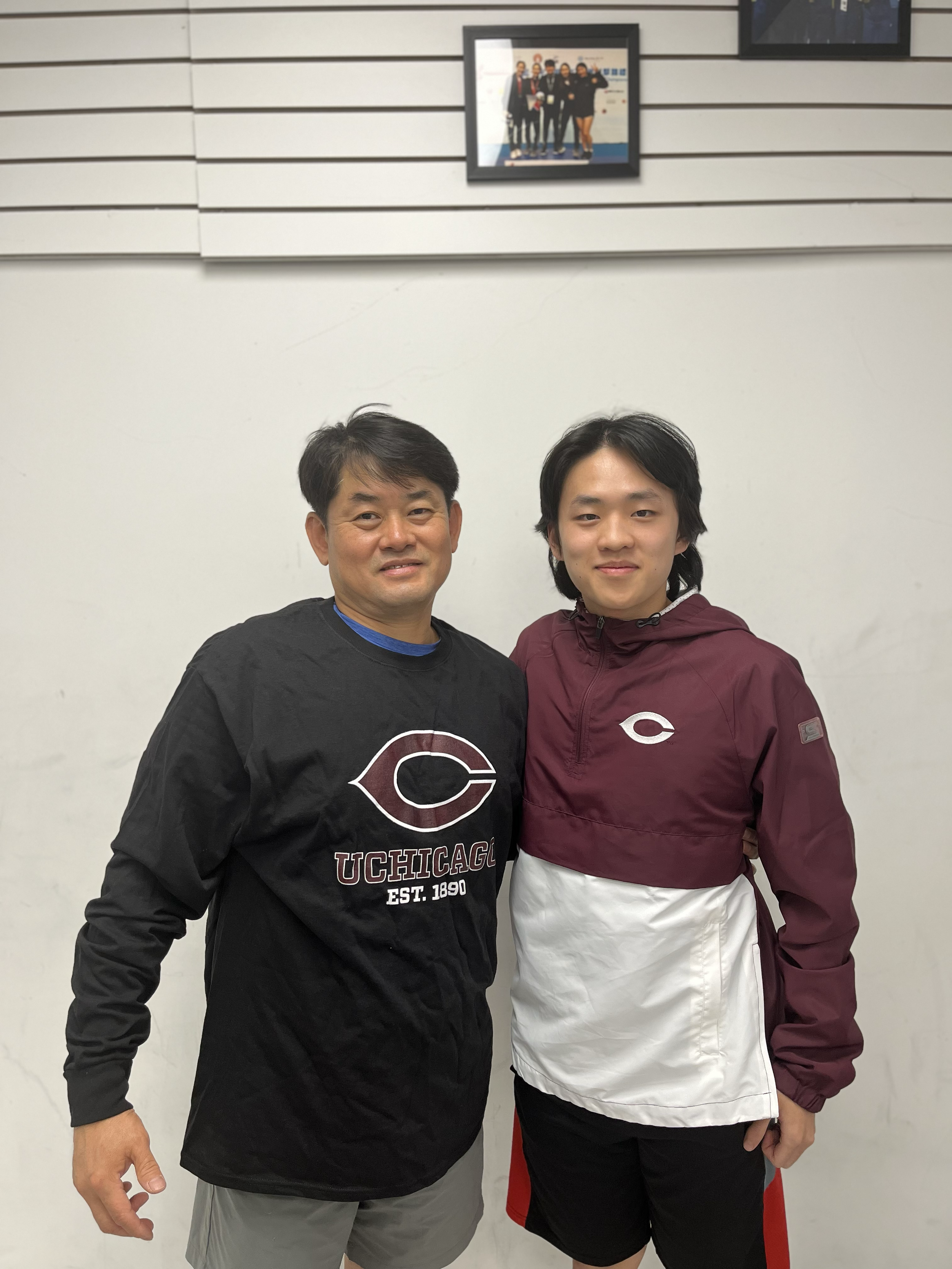 Two men standing side by side, smiling, in front of a plain white wall with a framed photo hanging above them. The man on the left is wearing a black sweatshirt with a Chicago Cubs logo and text. The man on the right is wearing a maroon and white jac