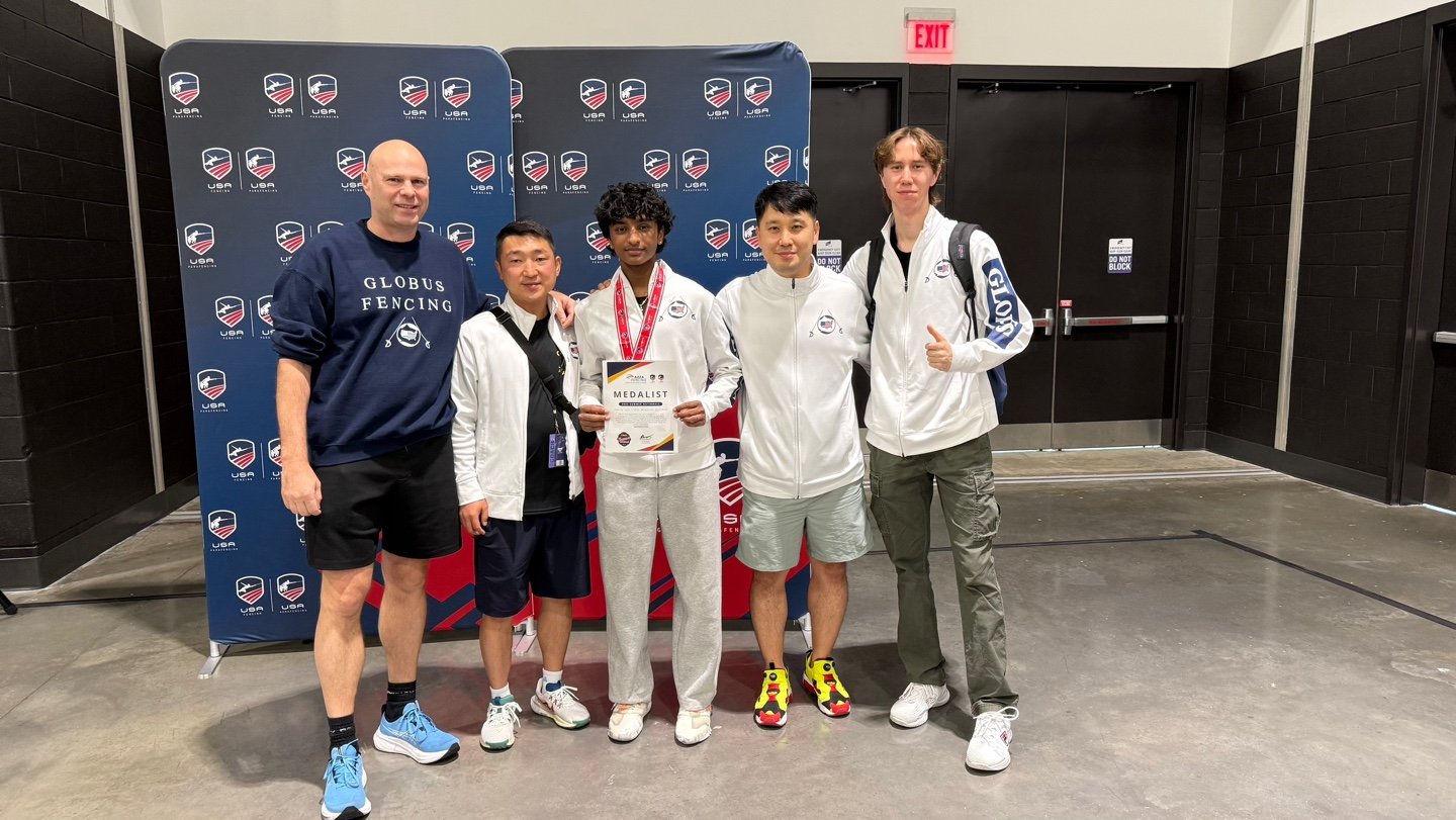 Group of five people standing in front of a USA Fencing backdrop at an indoor event, with one person holding a medal and certificate.