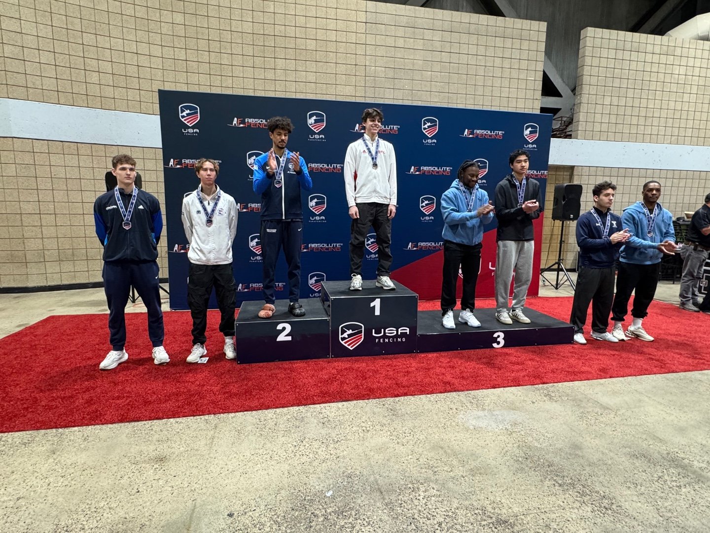 Young male athletes standing on a winners' podium at a fencing competition awards ceremony, with medals around their necks, in front of a blue backdrop with logos, and some clapping or looking forward.
