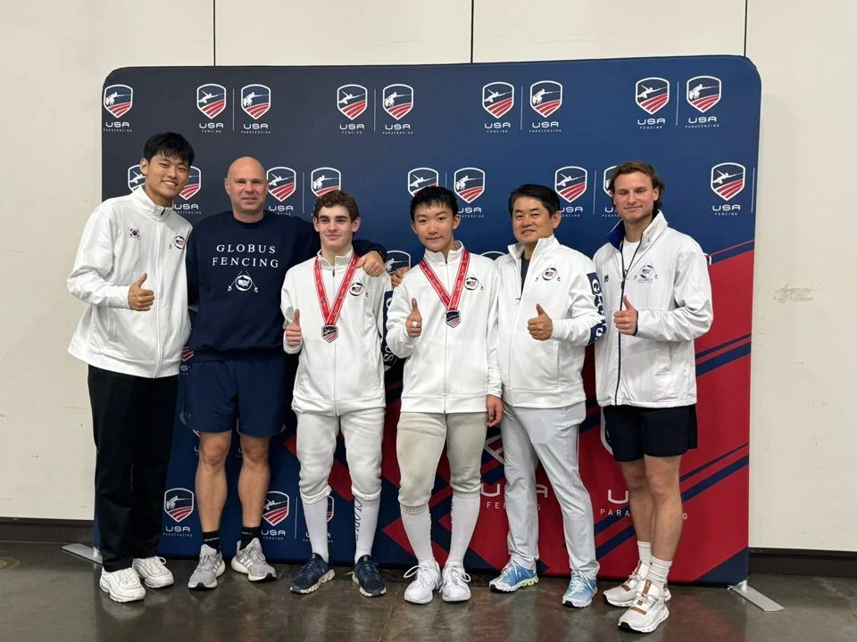 Group of six people standing in front of a USA fencing backdrop, three wearing medals, all giving thumbs up.