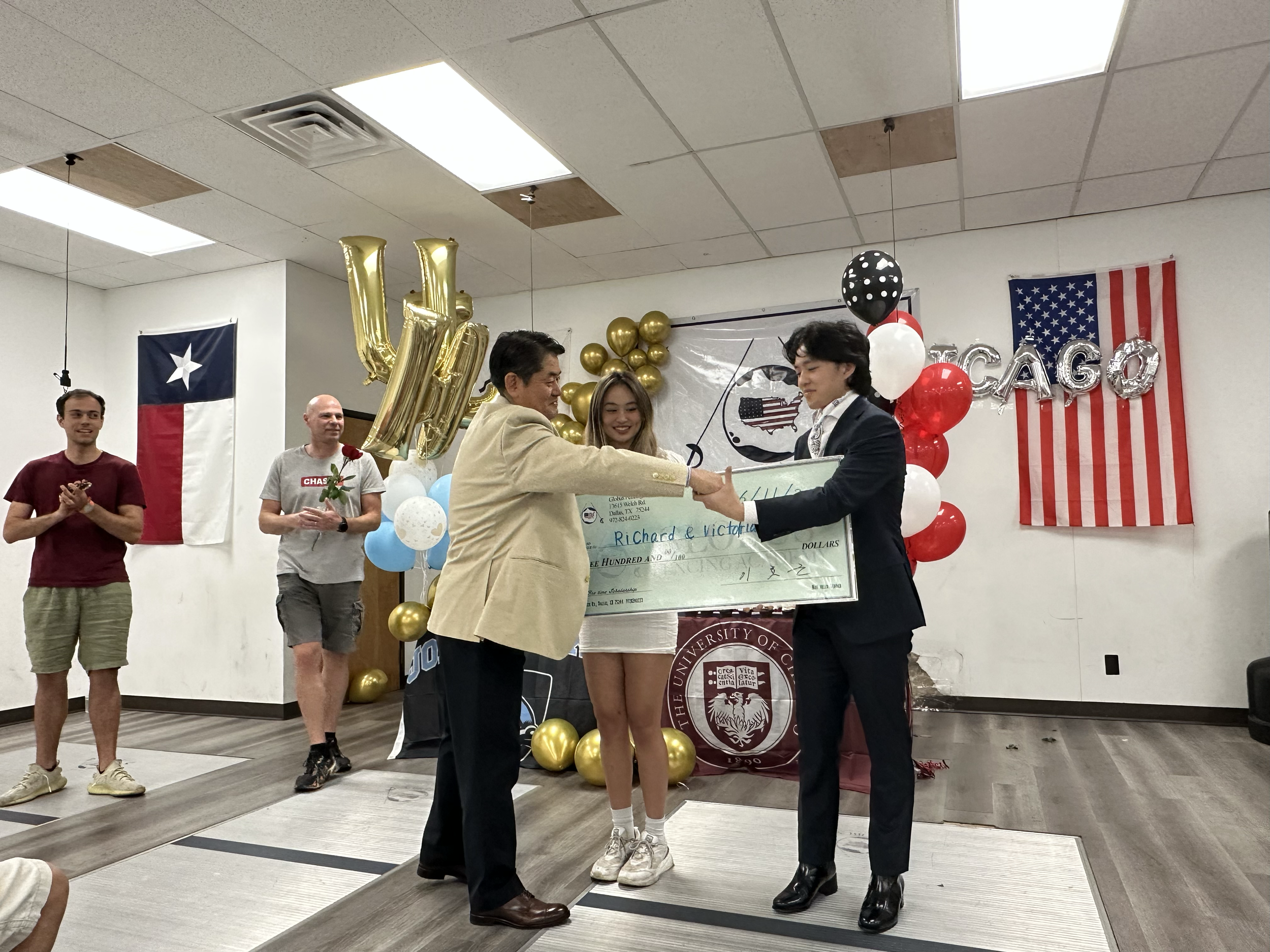 People celebrating at a graduation event with balloons, American and Texas flags, and a large check.
