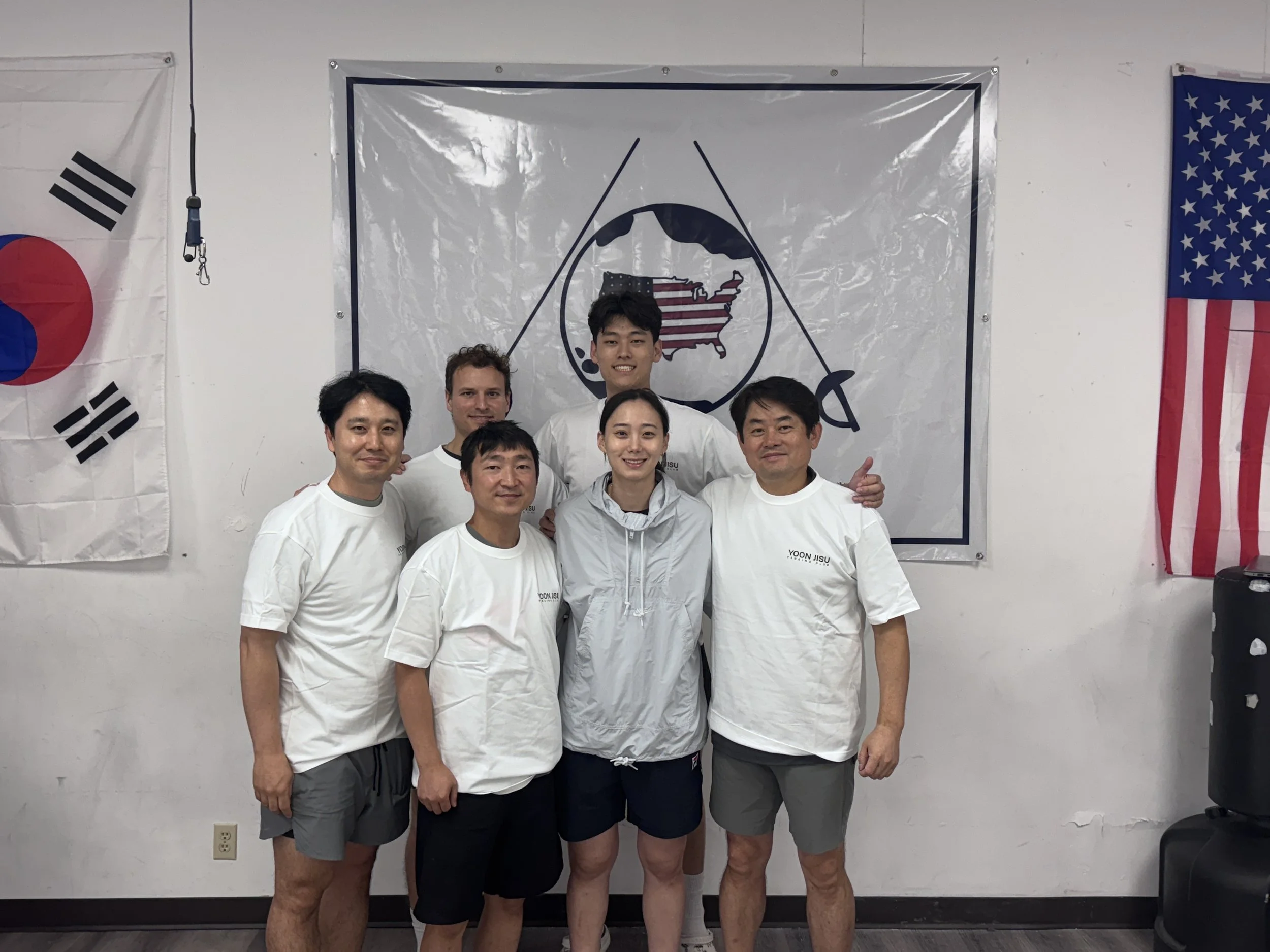 Group of six people in a martial arts dojo, wearing white martial arts uniforms, standing in front of flags of South Korea, the United States, and a martial arts emblem with a map of the United States and crossed swords.